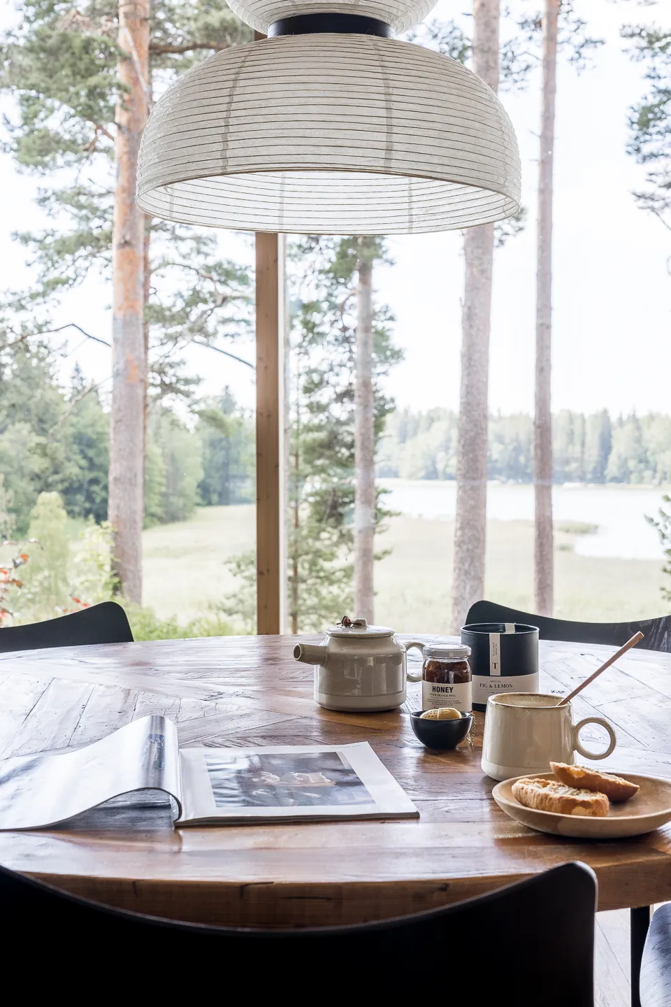 Pyöreällä puupöydällä on teepannu, hunajapurkki, teekuppi, leipää ja avattu aikakauslehti, ja suuri ikkuna avautuu metsämaisemaan.