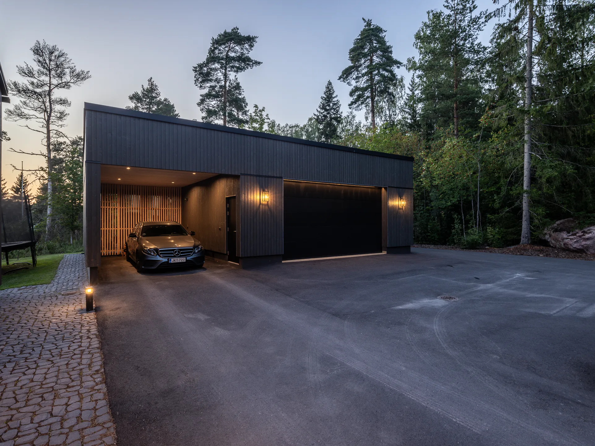 Moderni, tumma autotalli, jossa on harmaa auto, puuaita ja ulkovalot hämärässä.