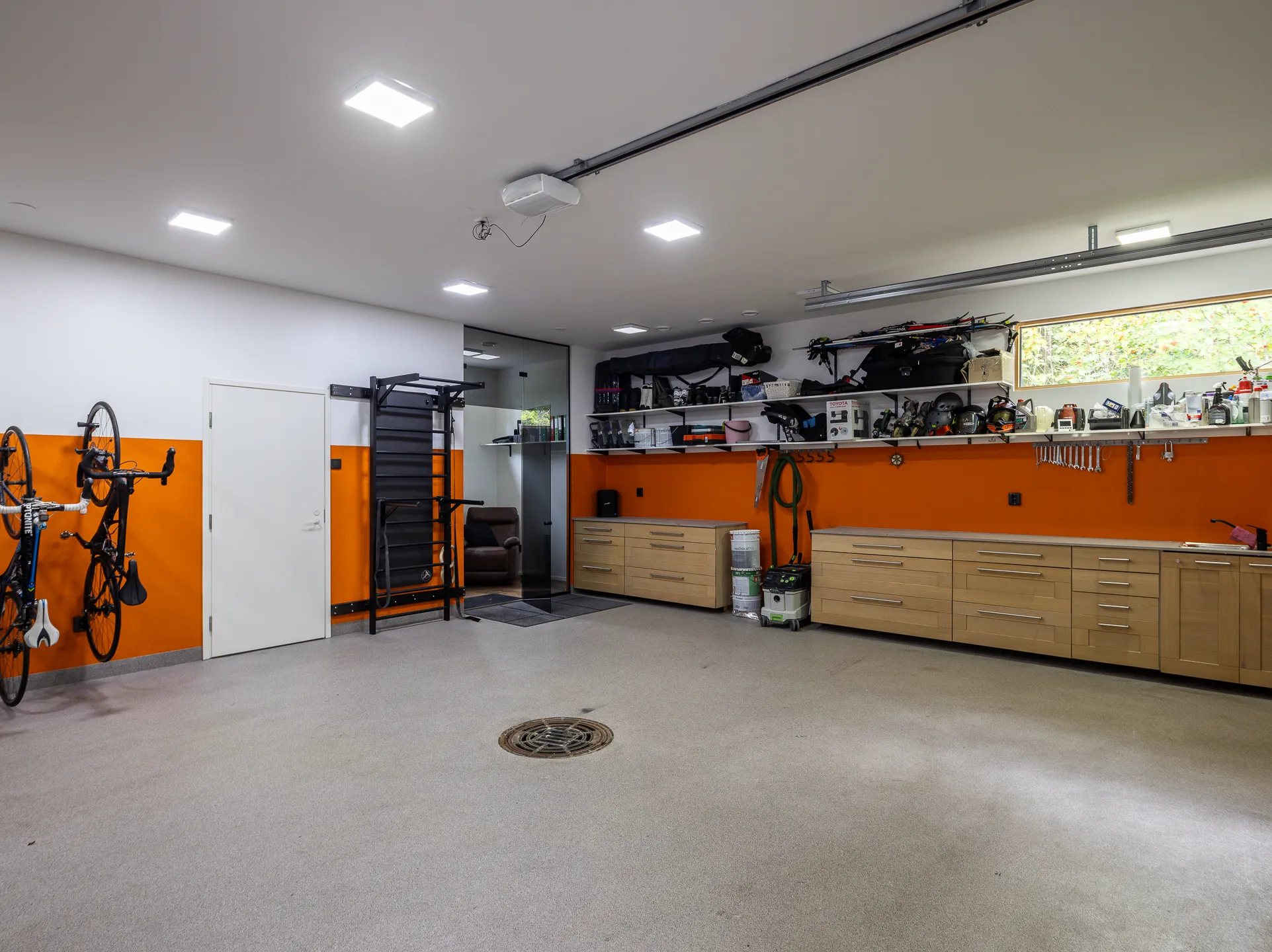Valoisassa autotallissa on oranssi seinä, vaaleat kaapit, polkupyöriä, treeniteline ja harmaa lattia.