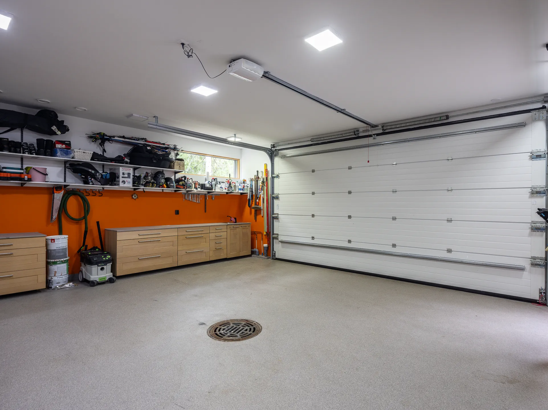 Valoisa autotalli, jossa on oranssi seinä, vaaleat puukaapit, valkoinen nosto-ovi ja harmaa lattia, sekä katossa valaisimet.