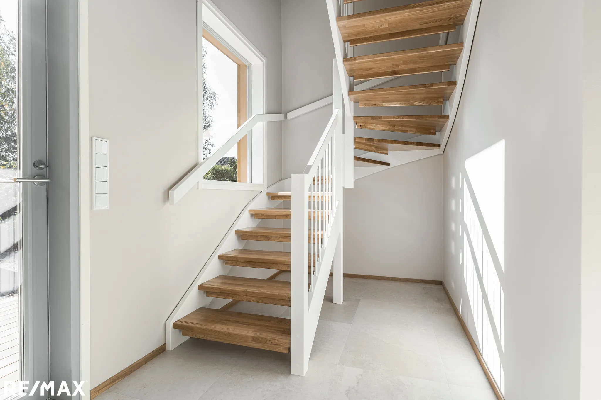 Vaaleassa eteisessä on puuportaat, valkoinen kaide ja suuri ikkuna, josta näkyy ulos. Lattialaatat ovat harmaat ja seinät vaaleat.