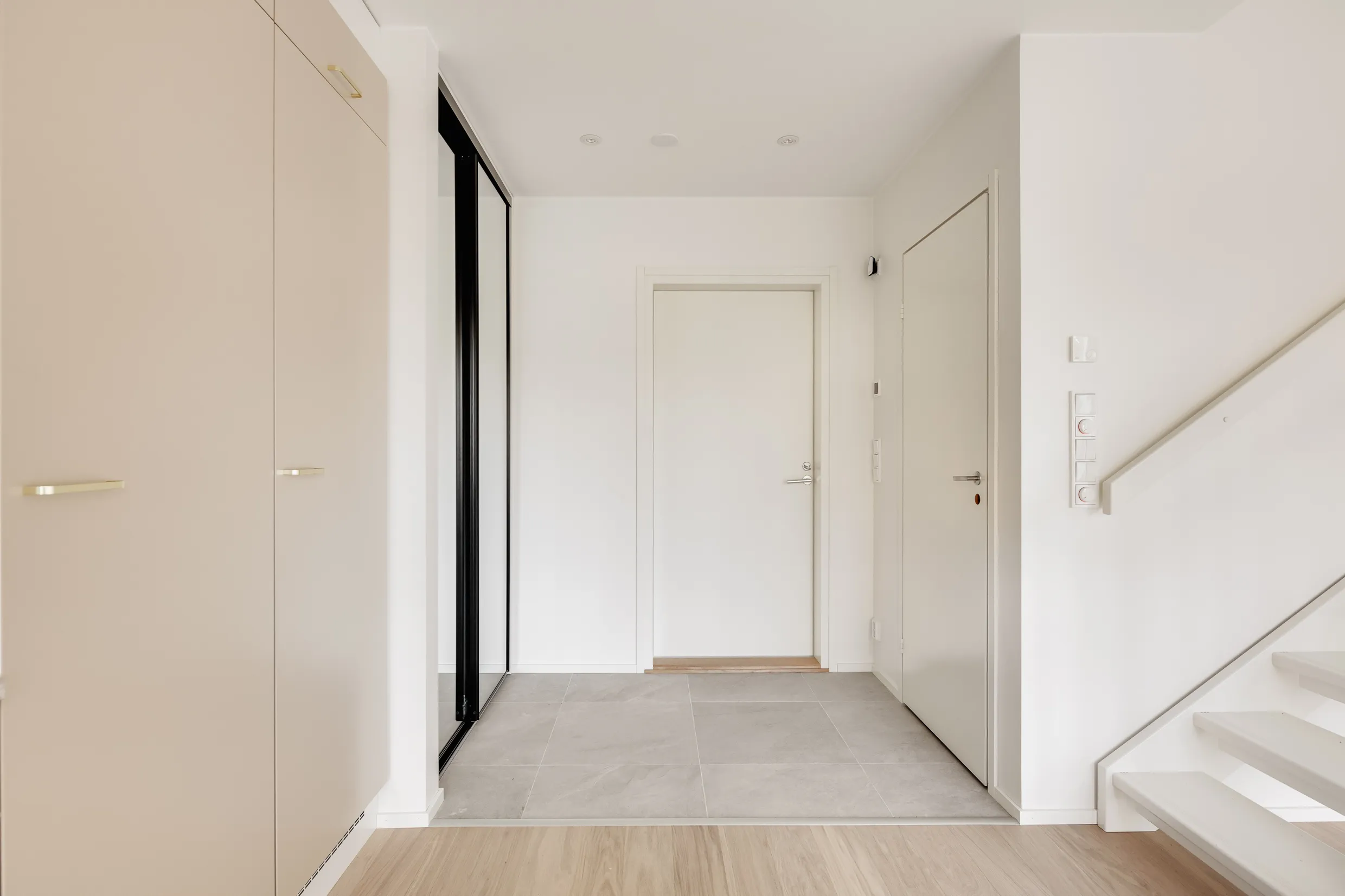 Vaalean eteisen sisustus, jossa on vaaleita kaappeja, peililiukuovi, valkoinen ovi ja portaat, sekä harmaa laattalattia ja vaalea puulattia.