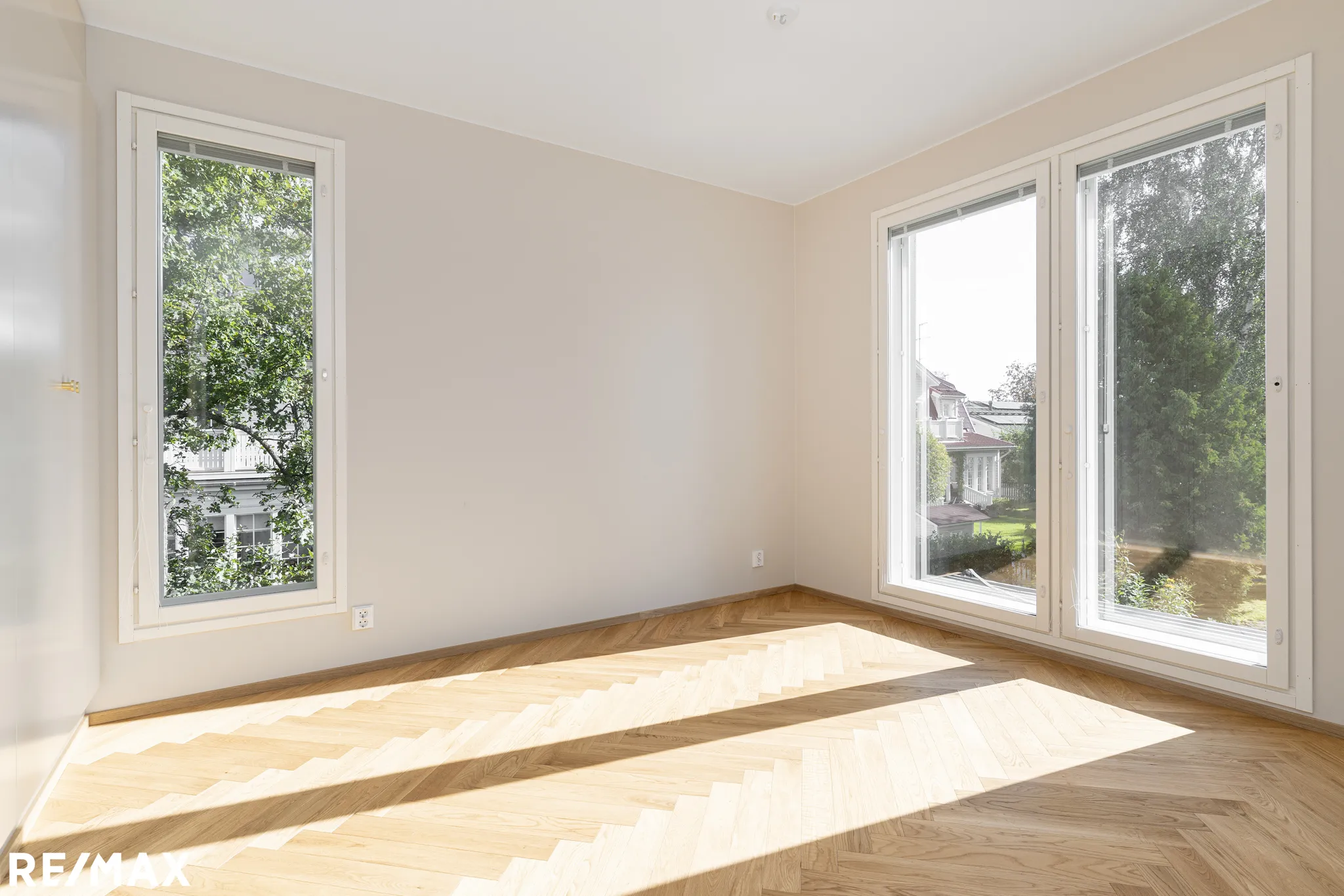 Valoisa huone, jossa on parkettilattia, kaksi ikkunaa ja valkoiset seinät. Auringonvalo lankeaa lattialle.