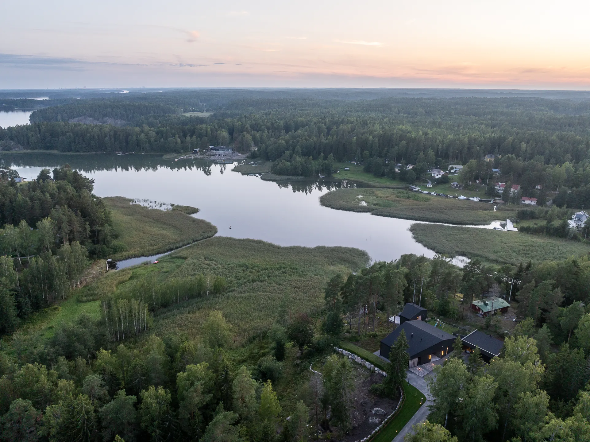 Ilmakuva järvimaisemasta auringonlaskun aikaan, jossa näkyy metsää, vettä ja muutamia rakennuksia. Taivaalla on vaaleanpunaisia ja oransseja sävyjä.
