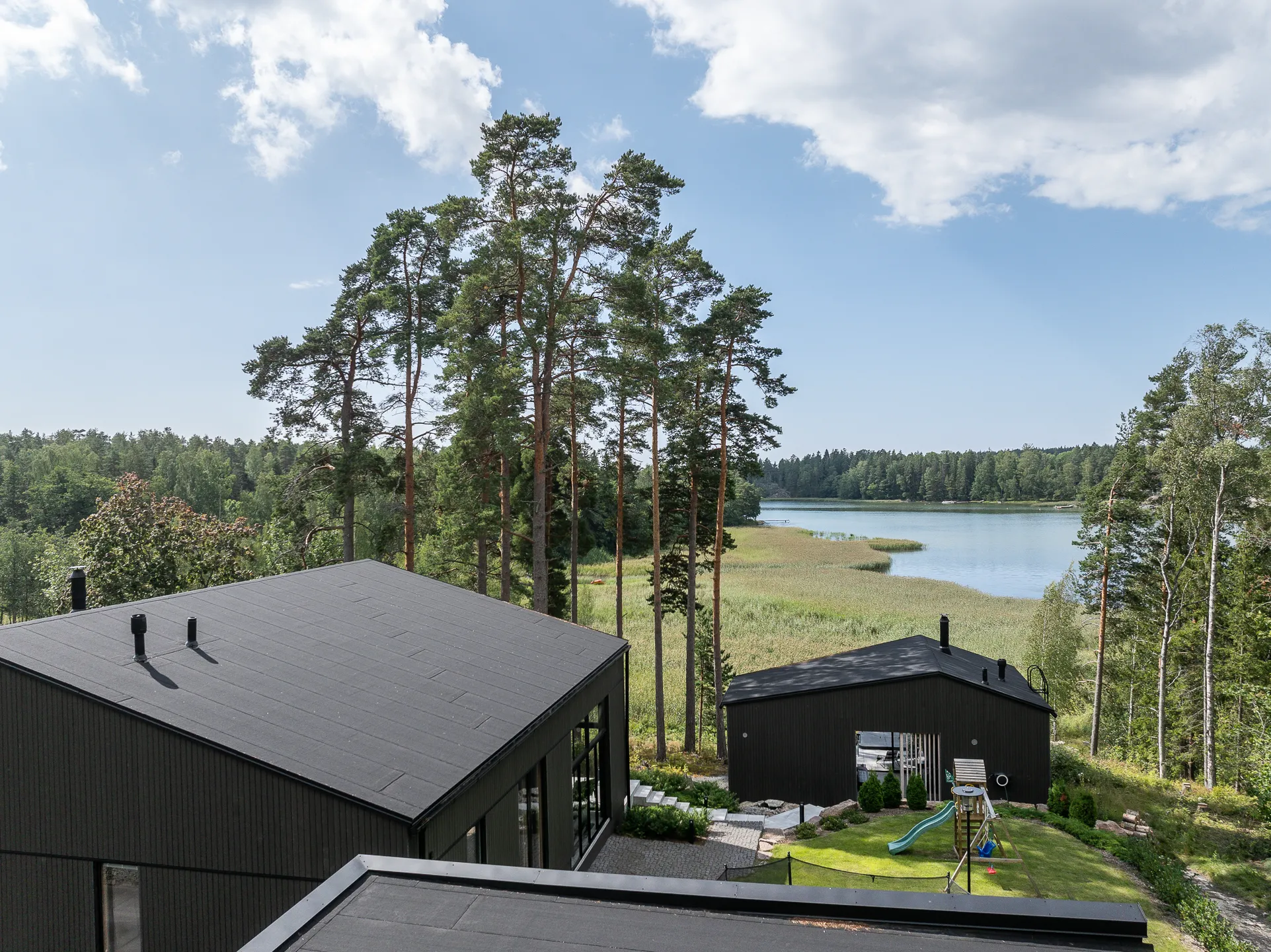 Panoraamanäkymä järvelle, jossa on tummat rakennukset, puut ja leikkipaikka aurinkoisena päivänä.