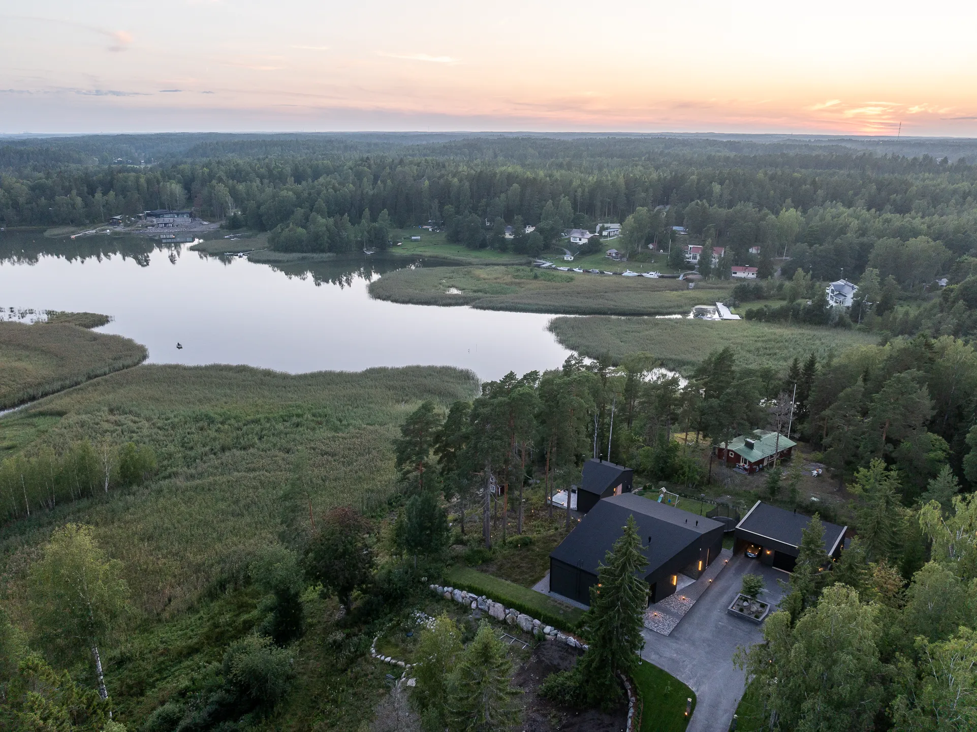 Ilmakuva järvimaisemasta auringonlaskun aikaan, jossa näkyy tumma talo, metsää ja vesistöä.