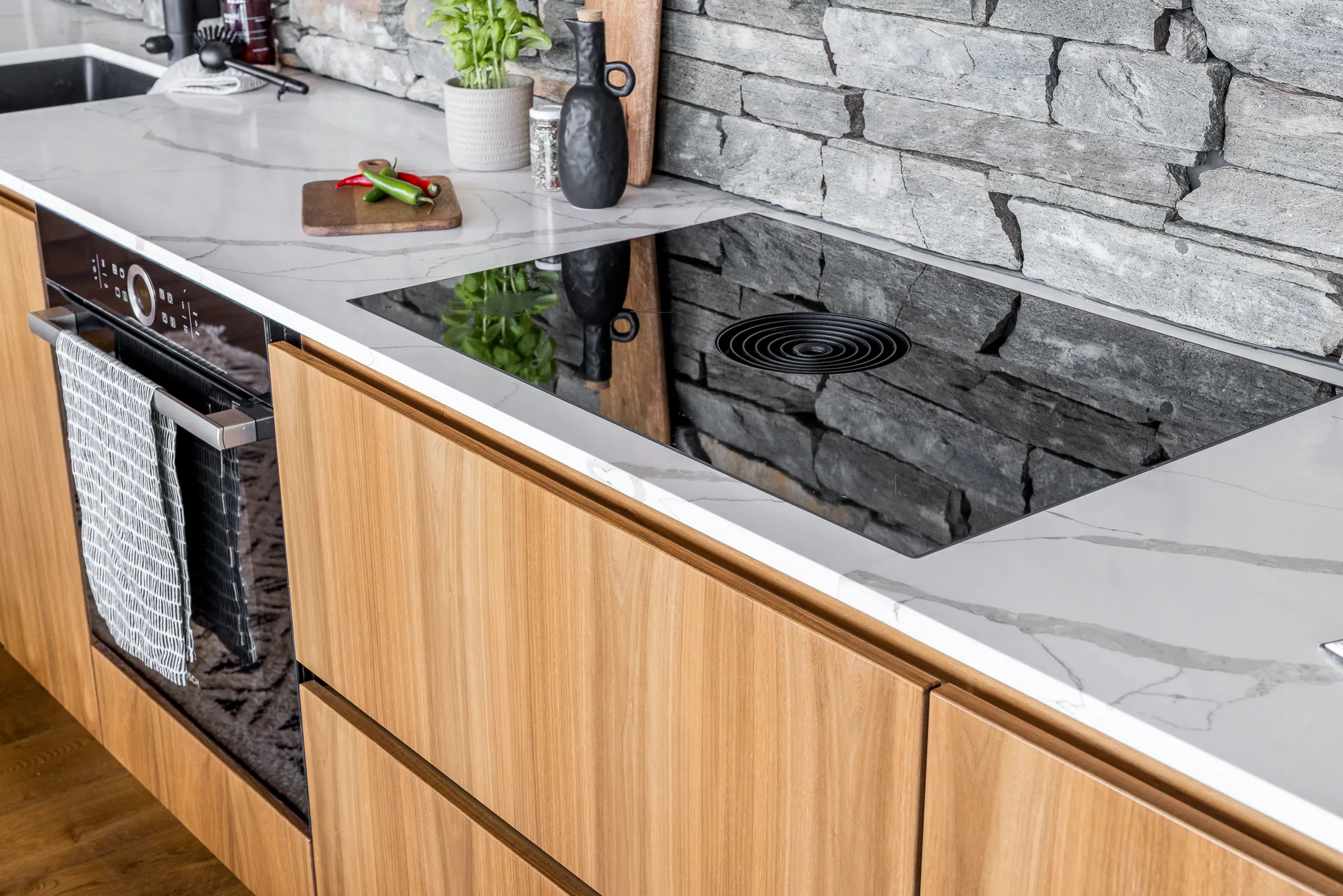 Moderni keittiö, jossa on puukaapit, marmorinen työtaso, induktioliesi ja kiviseinä. Työtasolla on myös pieni leikkuulauta, jossa on punaisia ja vihreitä chilejä.
