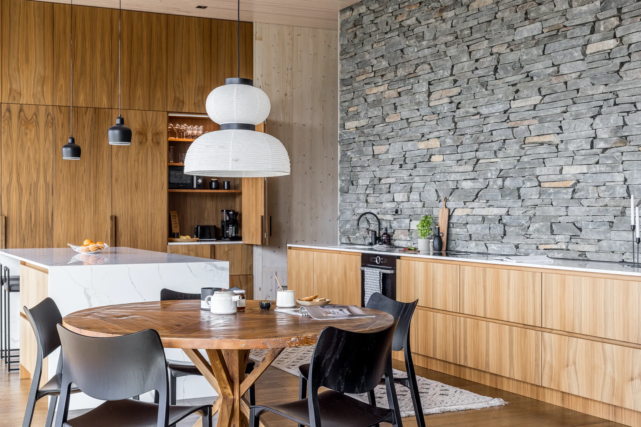 Moderni keittiö, jossa on puukaapit, kiviseinä, pyöreä puinen ruokapöytä ja mustat tuolit. Kaksi riippuvalaisinta valaisevat tilaa.