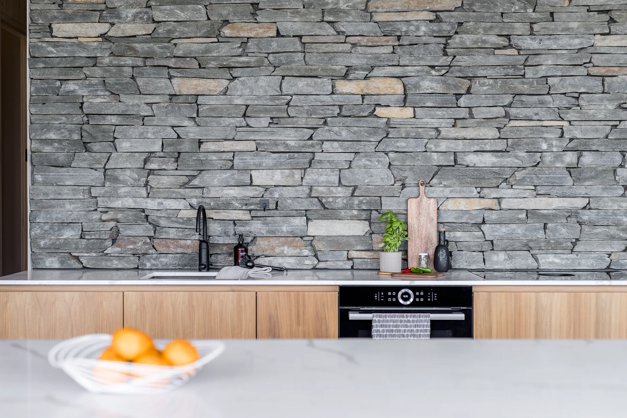 Keittiön kiviseinä, puukaapit, hedelmäkulho ja musta hana valoisassa tilassa.