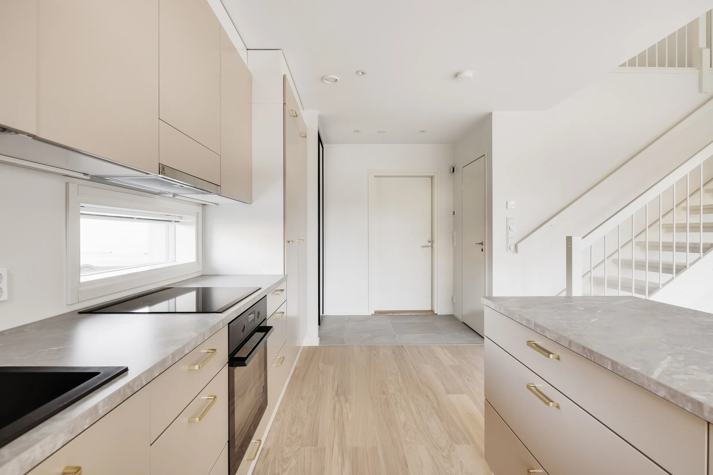 Vaaleassa keittiössä on vaaleat kaapit, harmaa työtaso, musta liesi ja uuni sekä valkoiset portaat. Lattia on vaaleaa puuta.