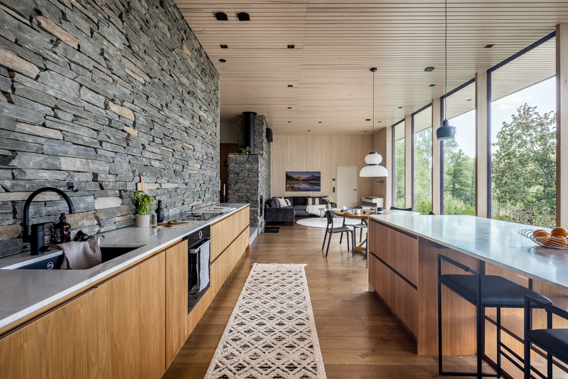 Moderni keittiö kiviseinällä, puukaapistoilla, saarekkeella ja suurilla ikkunoilla, joista näkyy metsä. Vaaleat puulattiat ja katto, musta hana ja valaisimet.