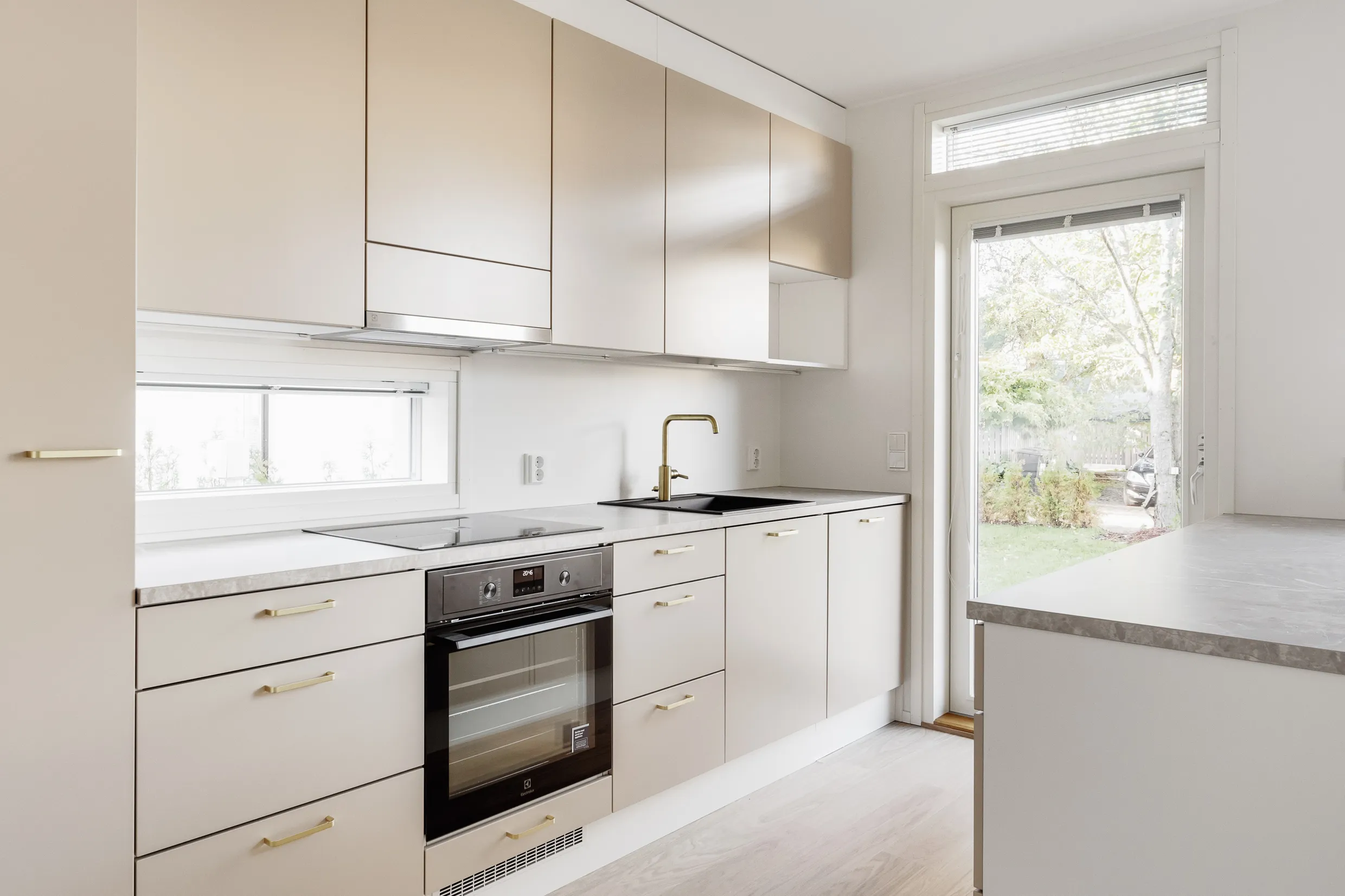 Vaaleassa keittiössä on beigejä kaappeja, kultainen hana ja musta pesuallas. Uuni on integroitu kaapistoon. Ikkunasta näkyy ulos.