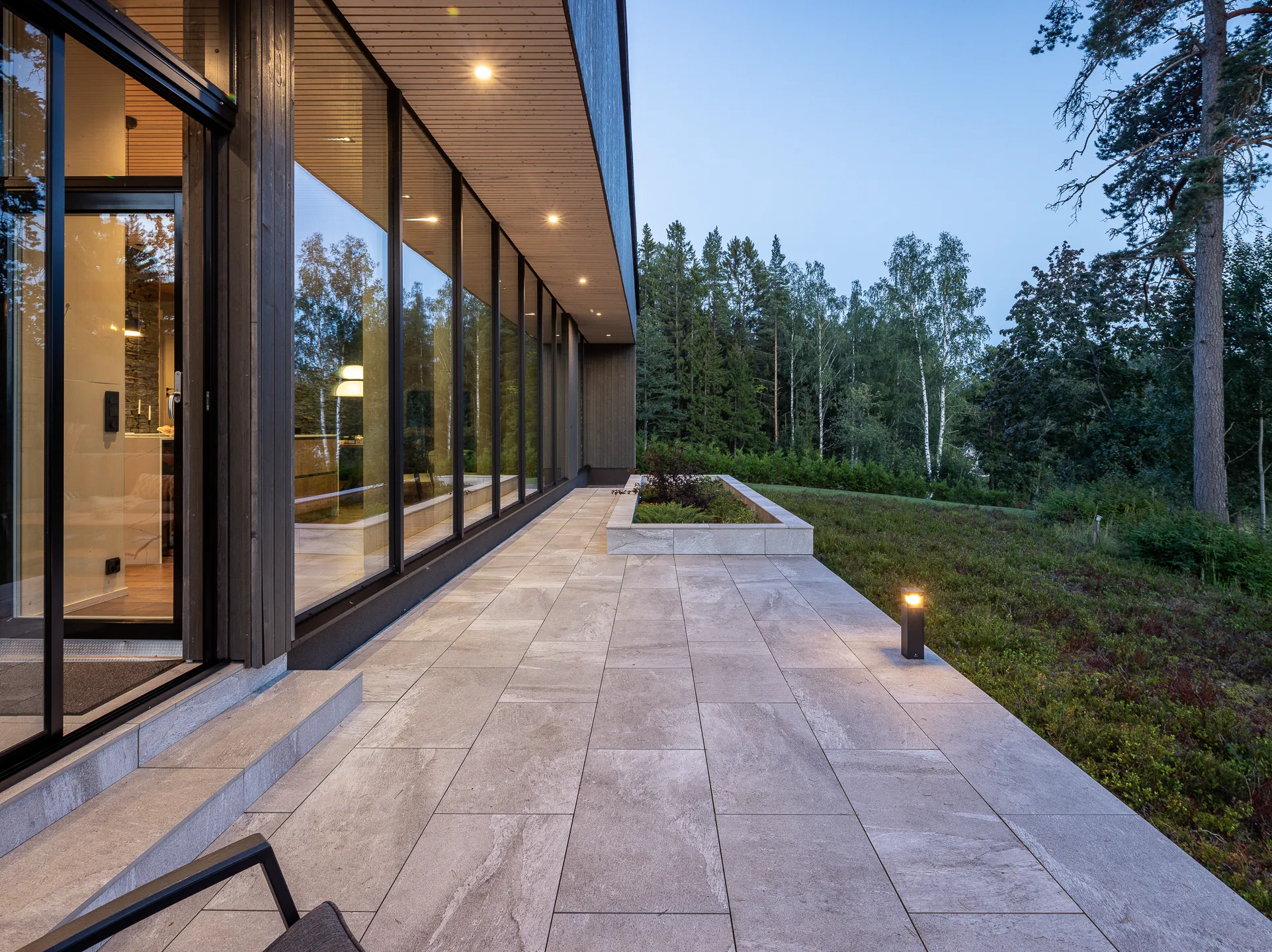Modernin talon kivetty terassi, jossa suuret ikkunat ja ulkovalaisin, metsä taustalla.