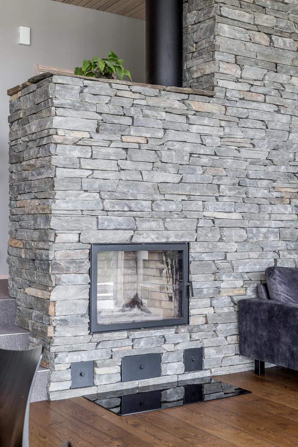 Kiviseinäinen takka, jossa on musta lasiluukku ja musta savupiippu, sekä musta suojalevy puulattialla.