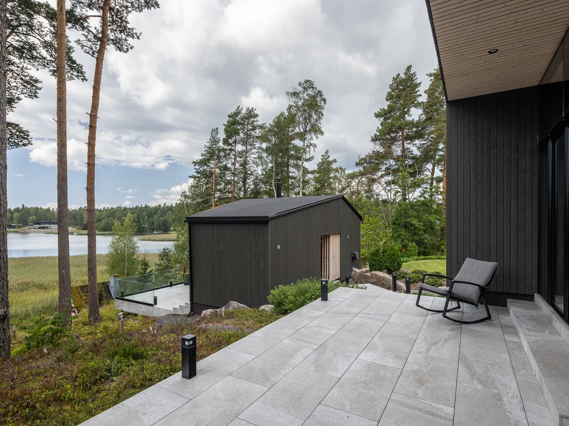 Ulkona on harmaa kiviterassi, jolla on tumma puurakennus, järvi ja metsä taustalla, sekä harmaa keinutuoli.