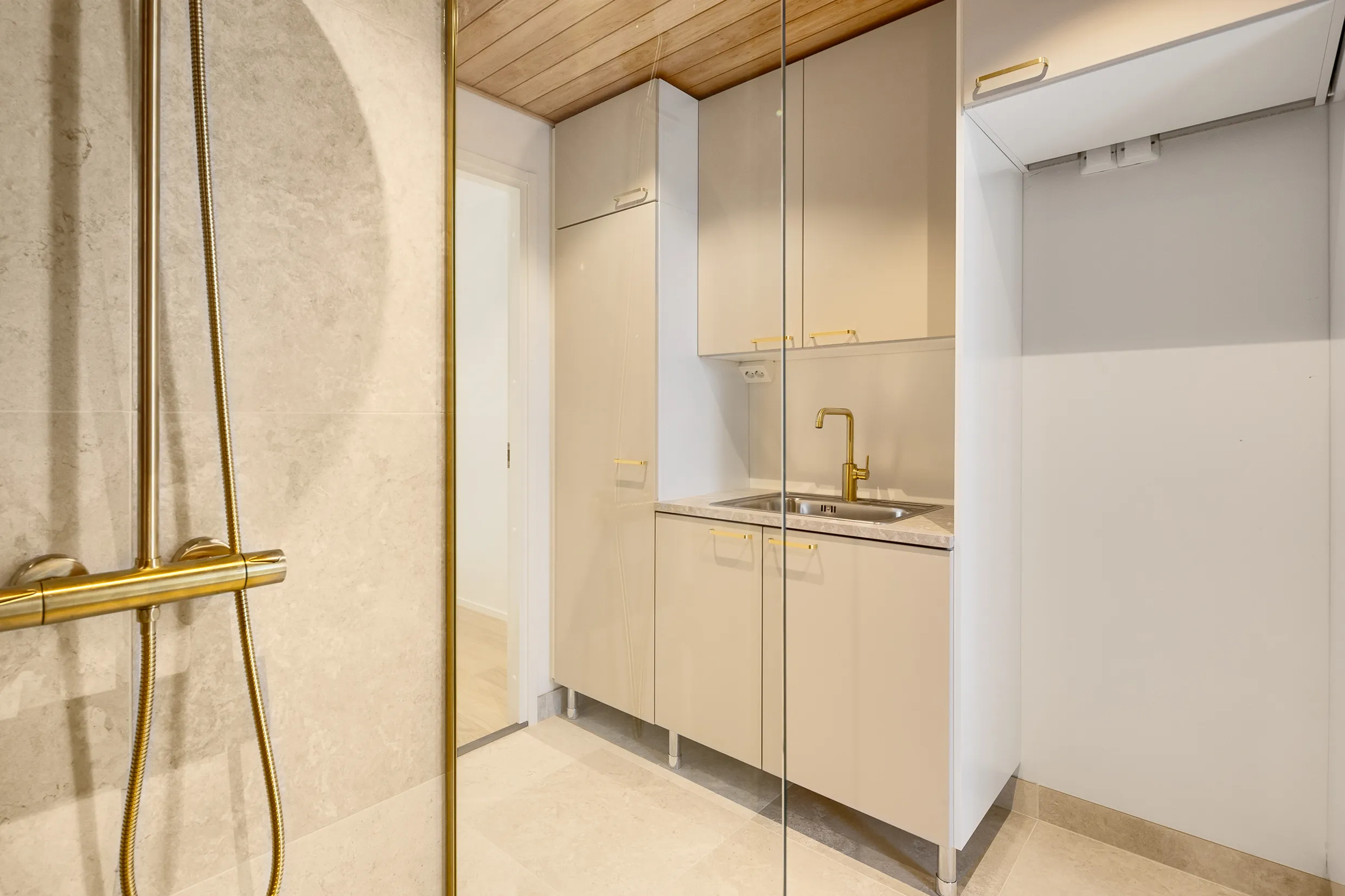 Kylpyhuoneen sisustus, jossa on beigejä kaappeja, kultainen hana ja suihku, sekä puinen katto ja marmorilattia.