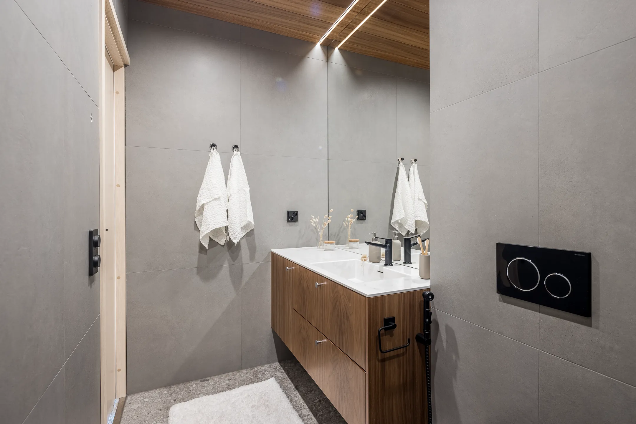 Moderni kylpyhuone, jossa on harmaat laatat, puinen katto ja pesuallas, valkoiset pyyhkeet ja musta wc-painike.