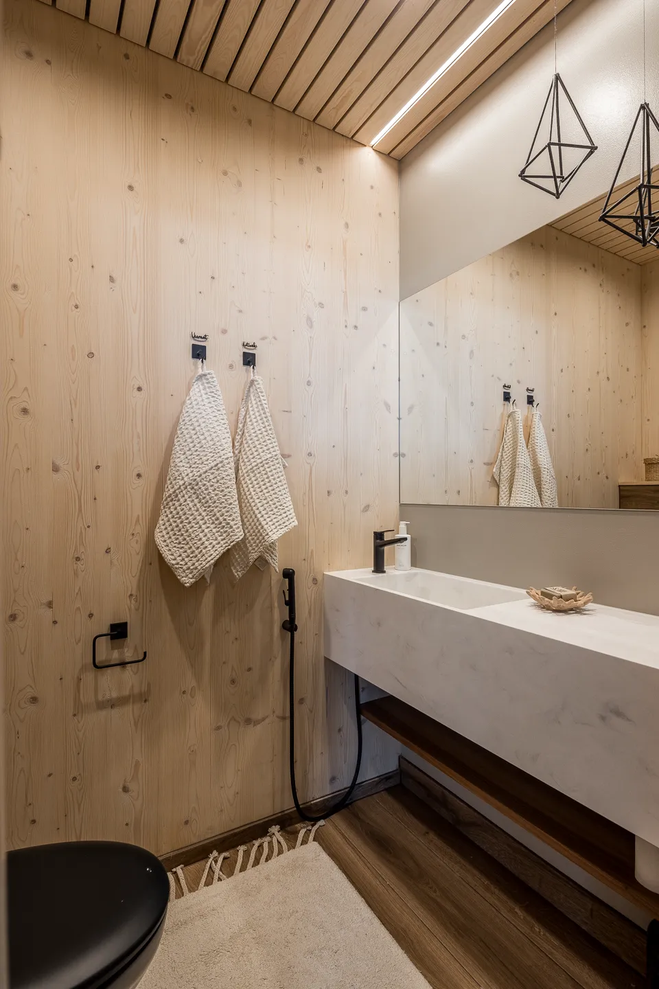 Vaalean puun sävyinen kylpyhuone, jossa on valkoinen pesuallas, musta hana ja kaksi pyyhettä seinällä.