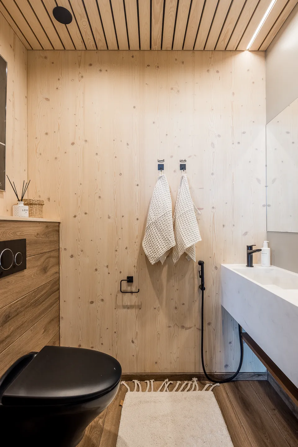 Moderni kylpyhuone, jossa puupanelointi, musta WC-istuin, valkoinen pesuallas ja kaksi valkoista pyyhettä roikkuu seinällä.
