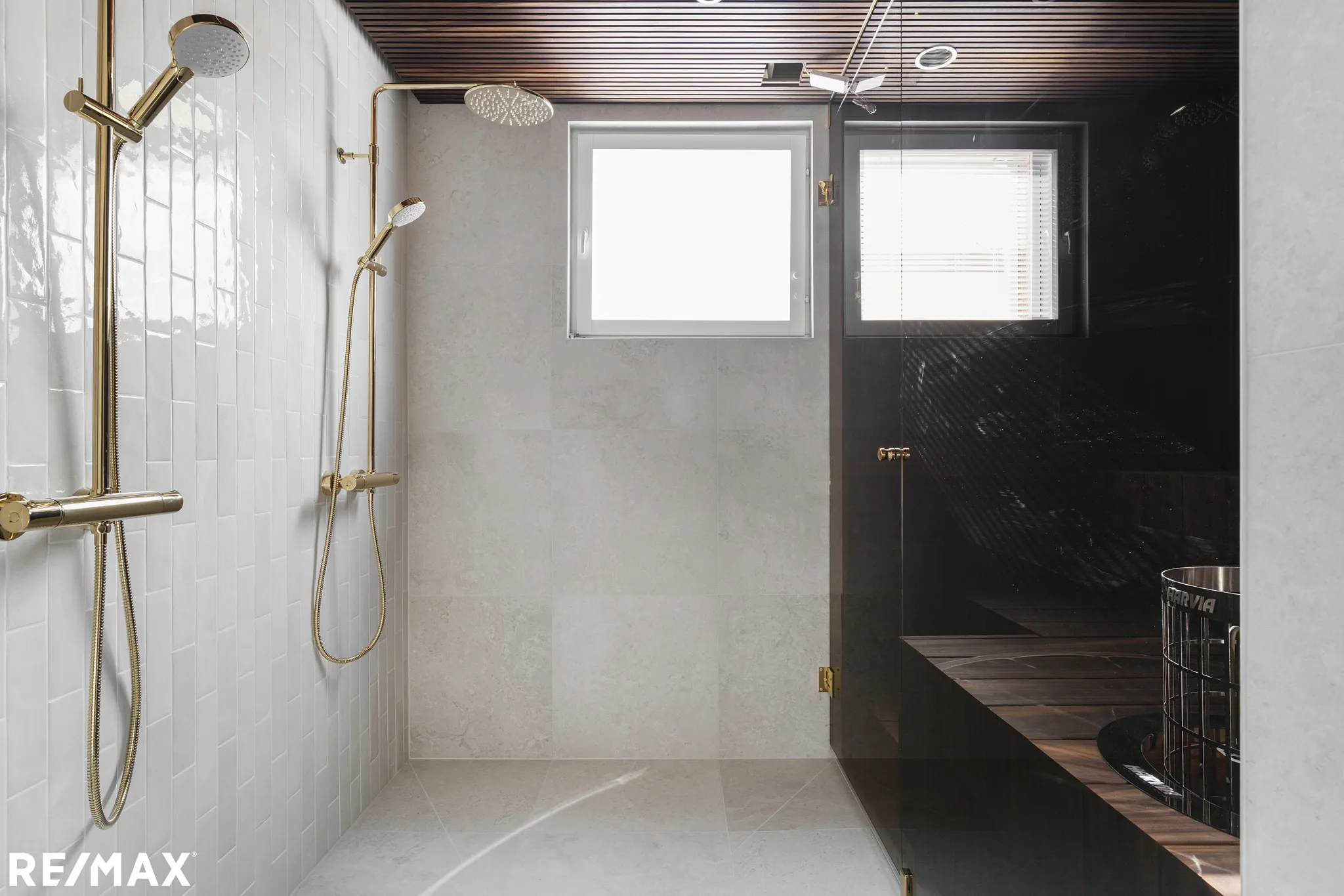 Kylpyhuoneen sisustus, jossa on kultaiset suihkut, valkoiset ja harmaat laatat, puinen sauna ja ikkunat.