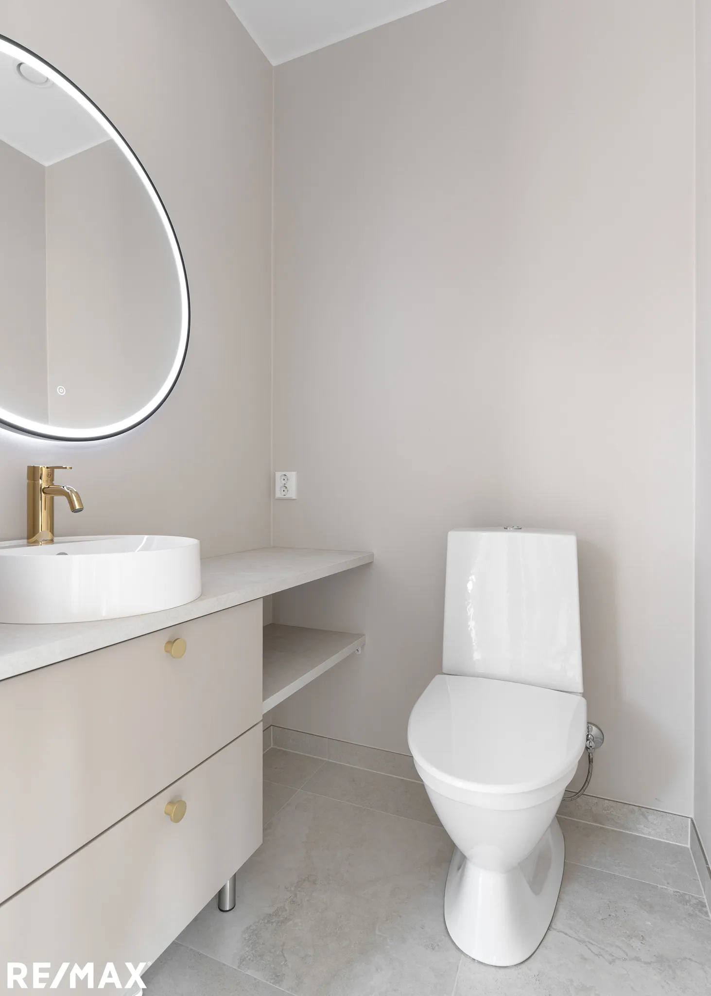 Valoisa kylpyhuone, jossa on pyöreä peili, valkoinen pesuallas, beige lipasto, valkoinen wc-istuin ja harmaat laatat.