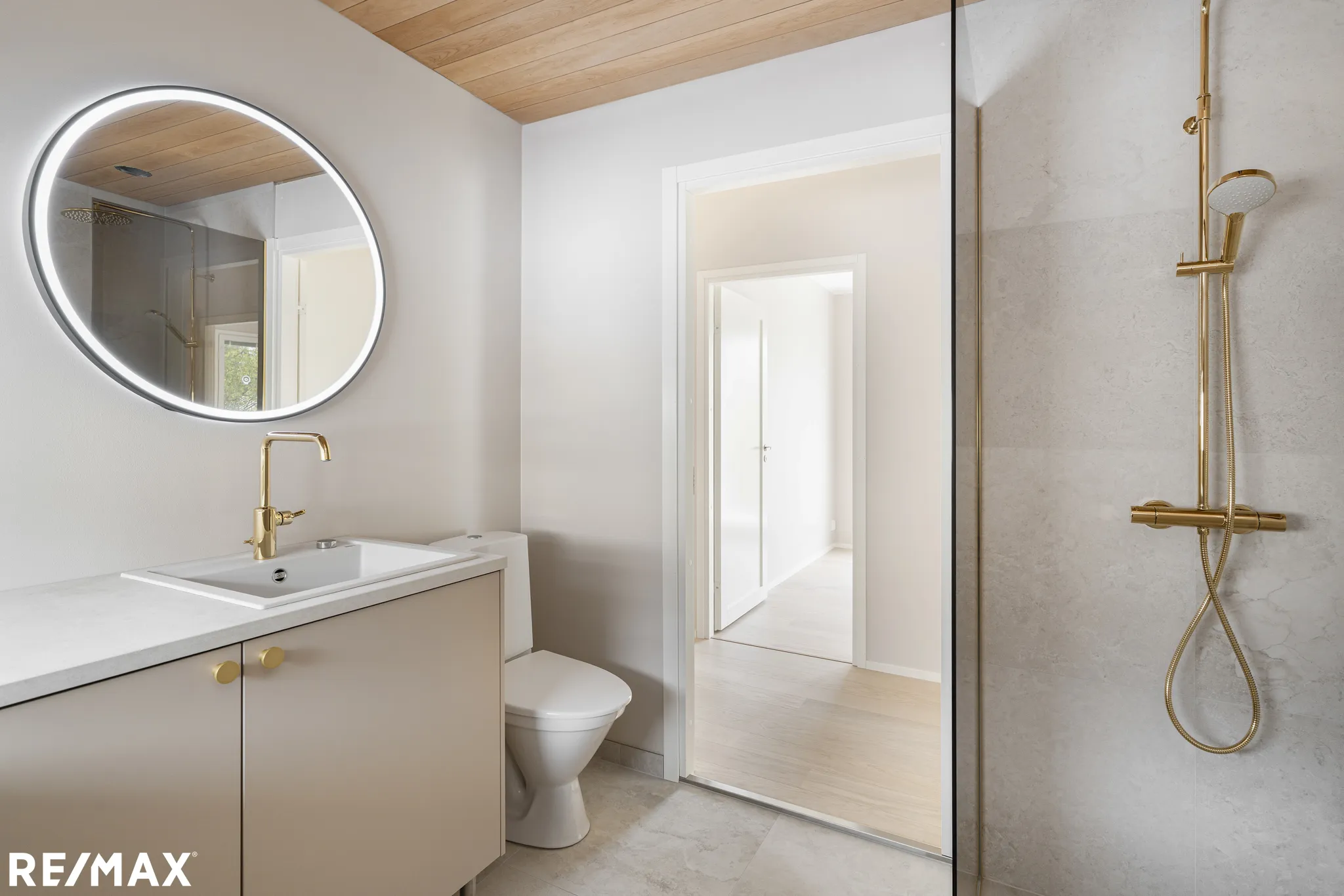 Vaaleassa kylpyhuoneessa on pyöreä peili, beige allaskaappi, valkoinen wc-istuin ja kultainen suihku. Puinen kattopaneeli ja avoin oviaukko näkyvät.