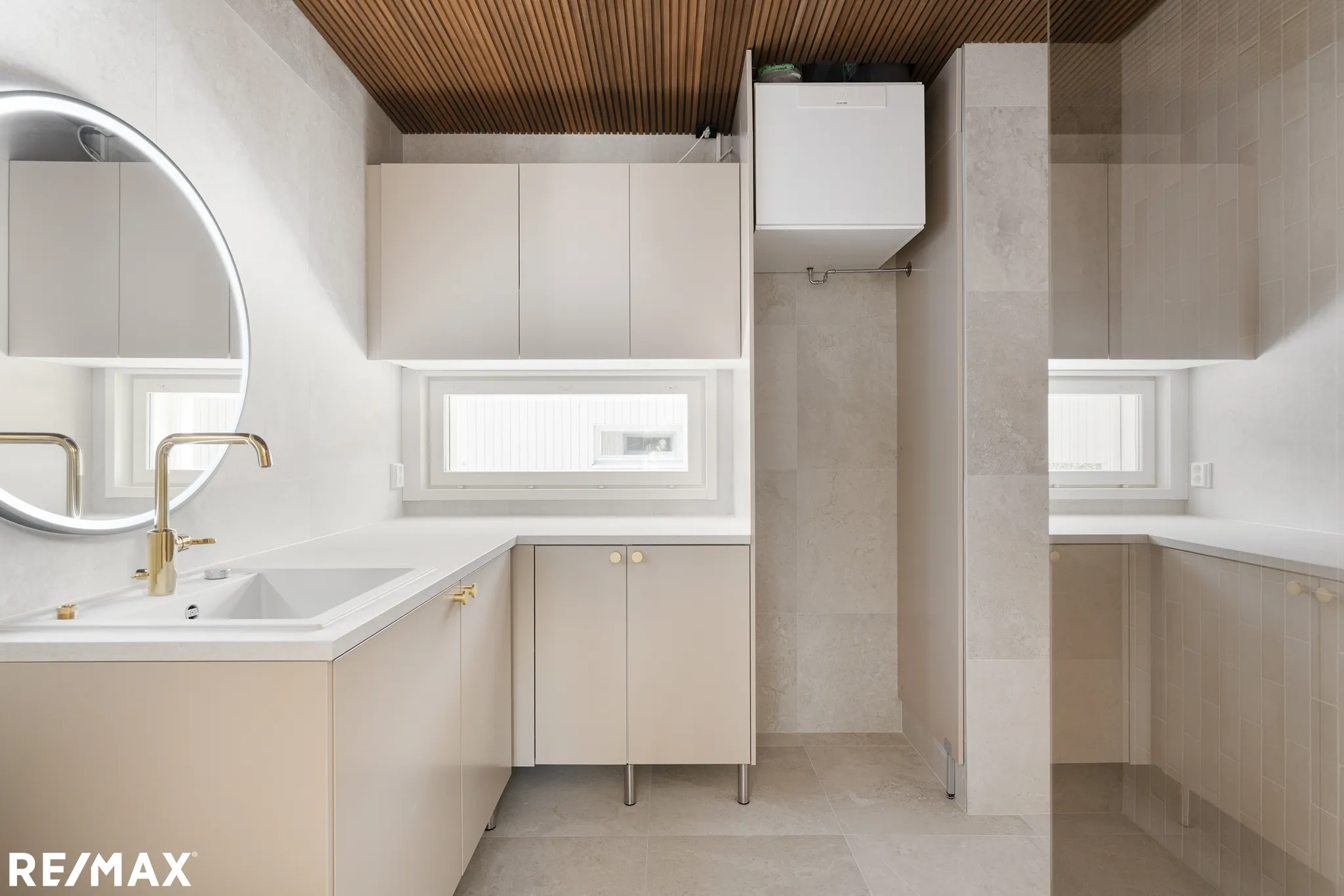 Vaalean sävyinen kylpyhuone, jossa on pyöreä peili, valkoinen pesuallas, kultainen hana, puinen kattopaneeli ja vaaleat kaapit.