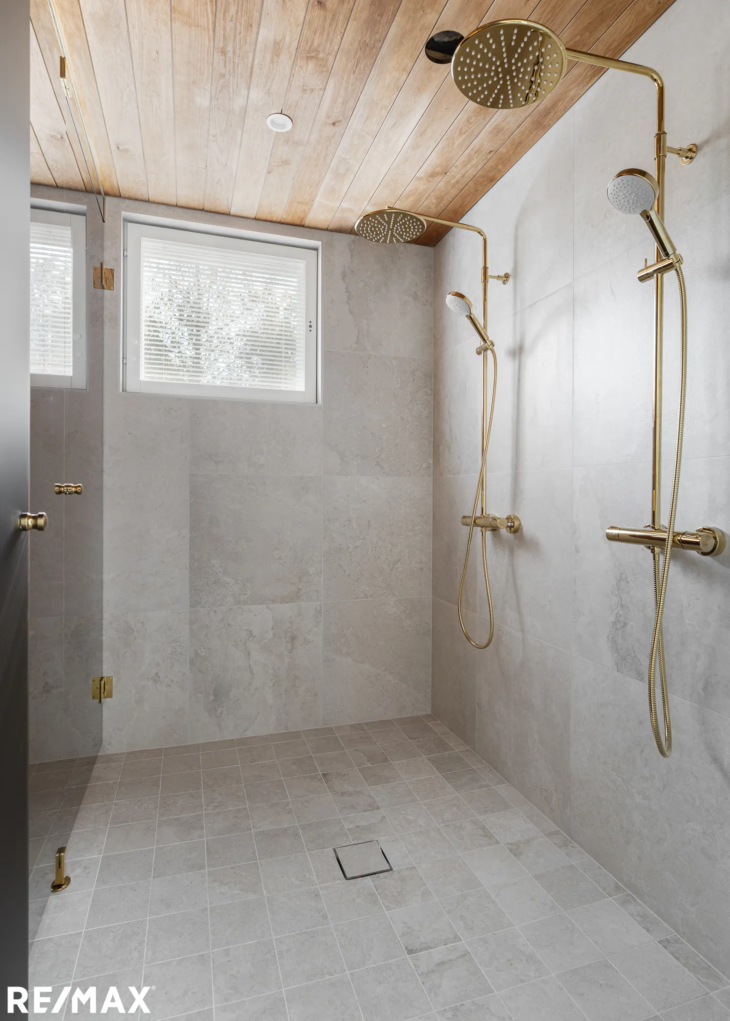 Kylpyhuoneen suihkussa on kultaiset suihkupäät, harmaat laatat ja puinen katto.