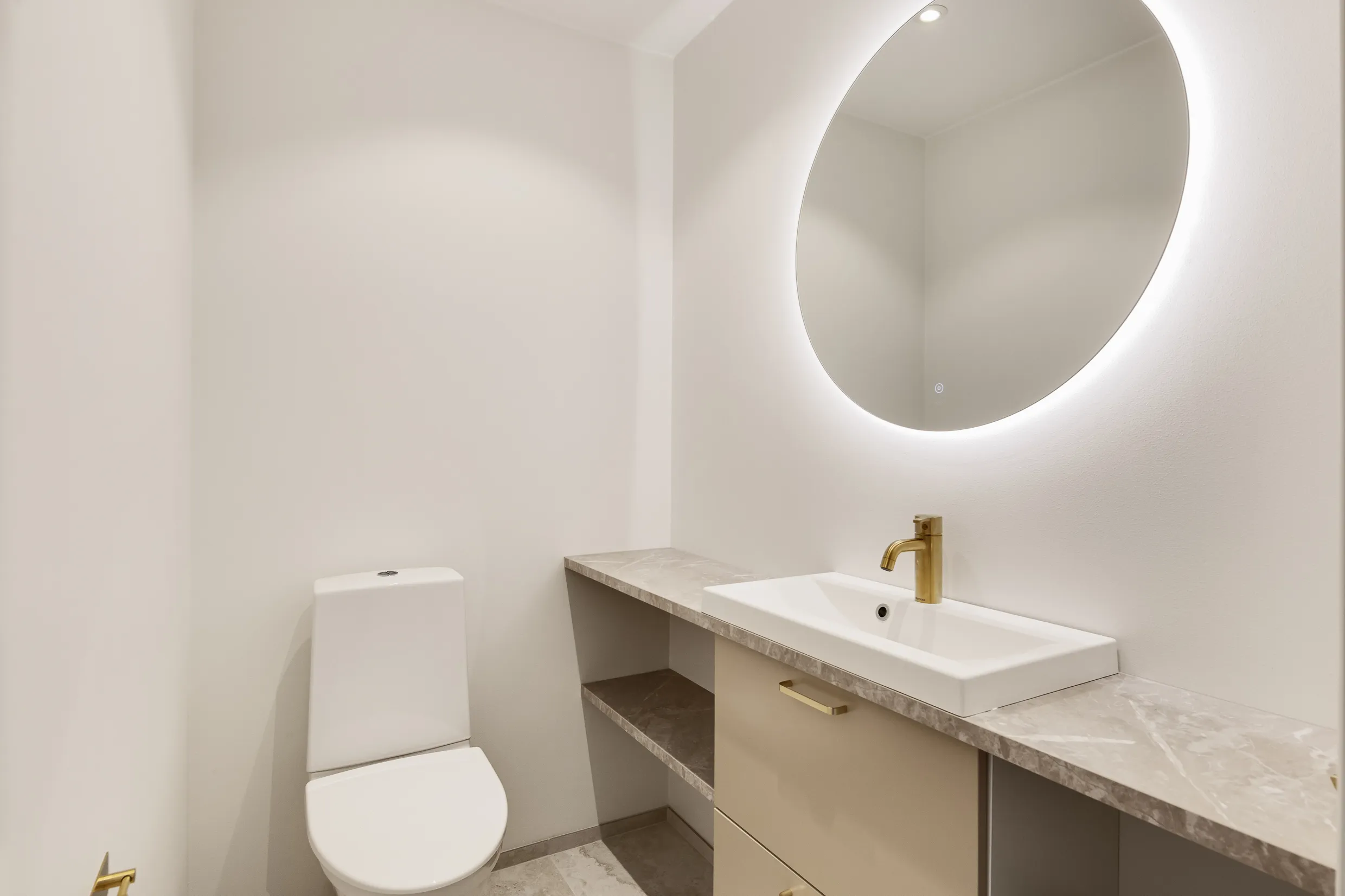 Vaaleassa kylpyhuoneessa on valkoinen wc-istuin, beige pesuallas kultaisella hanalla ja pyöreä peili, jossa on valaistus.