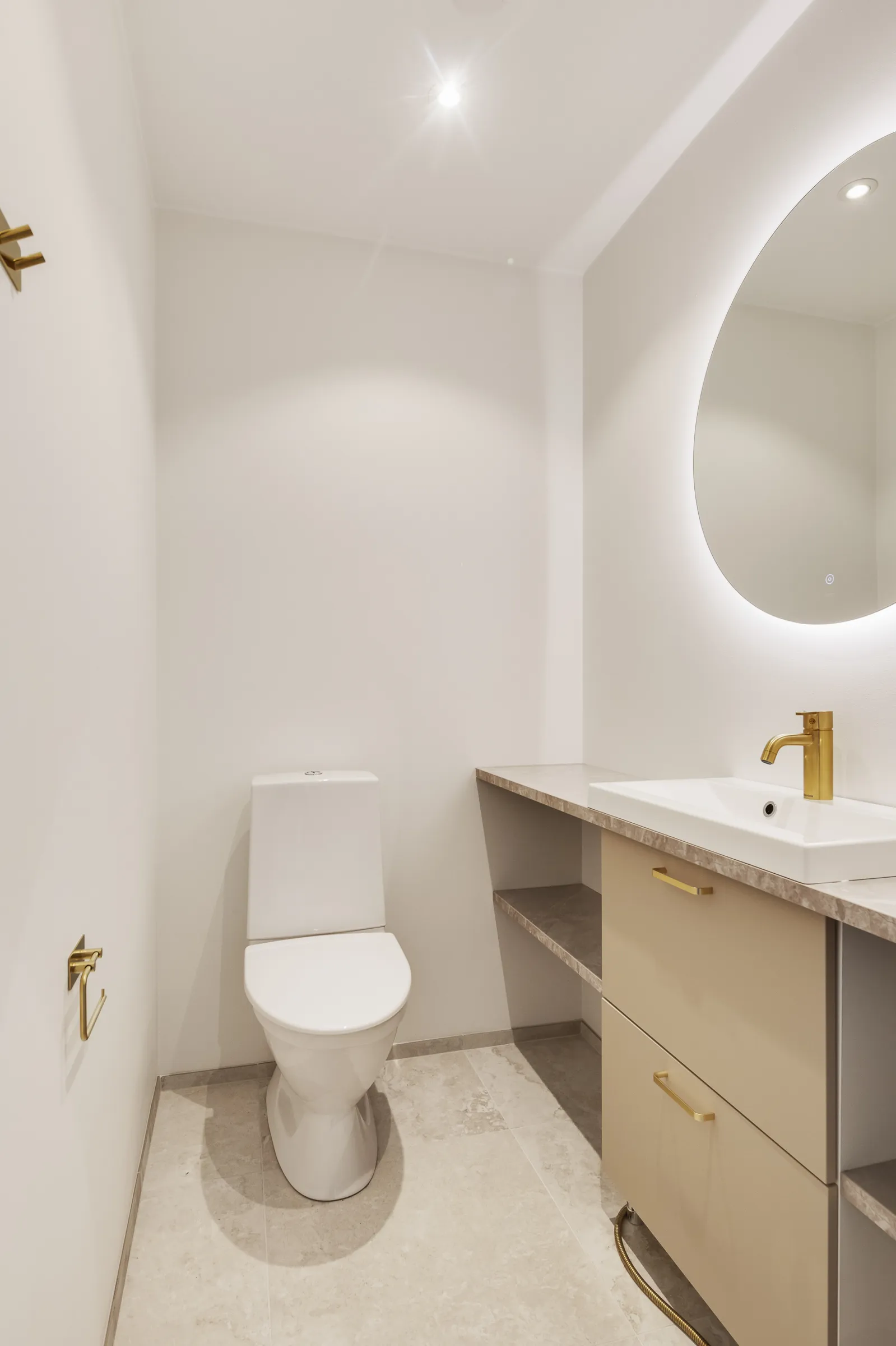 Vaaleassa kylpyhuoneessa on valkoinen wc-istuin, pyöreä peili valaistuksella, beige pesuallas kaapilla ja kultaiset yksityiskohdat.