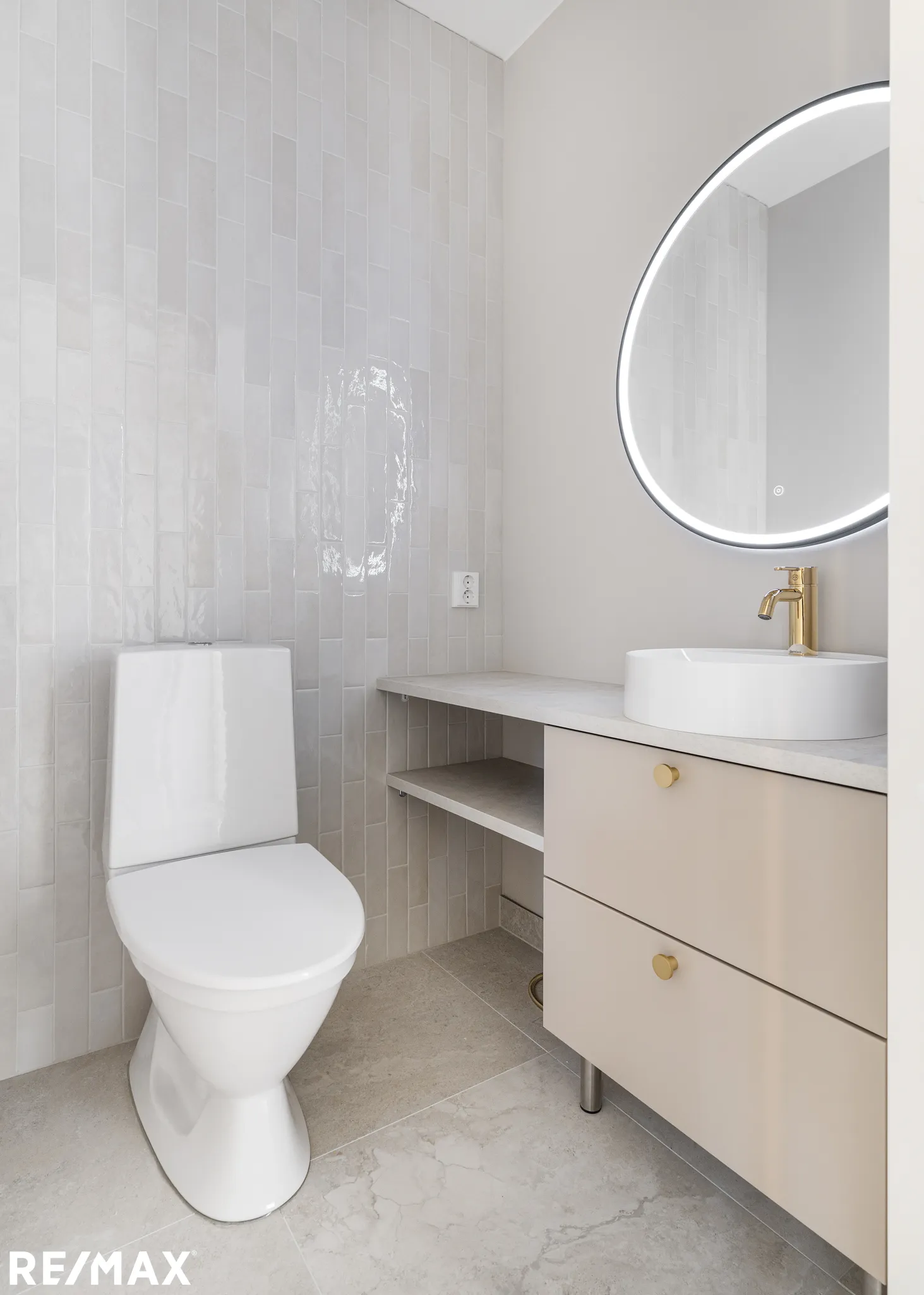 Vaaleassa kylpyhuoneessa on valkoinen wc-istuin, pyöreä peili valolla, beige pesuallaskaappi ja marmorilattia.