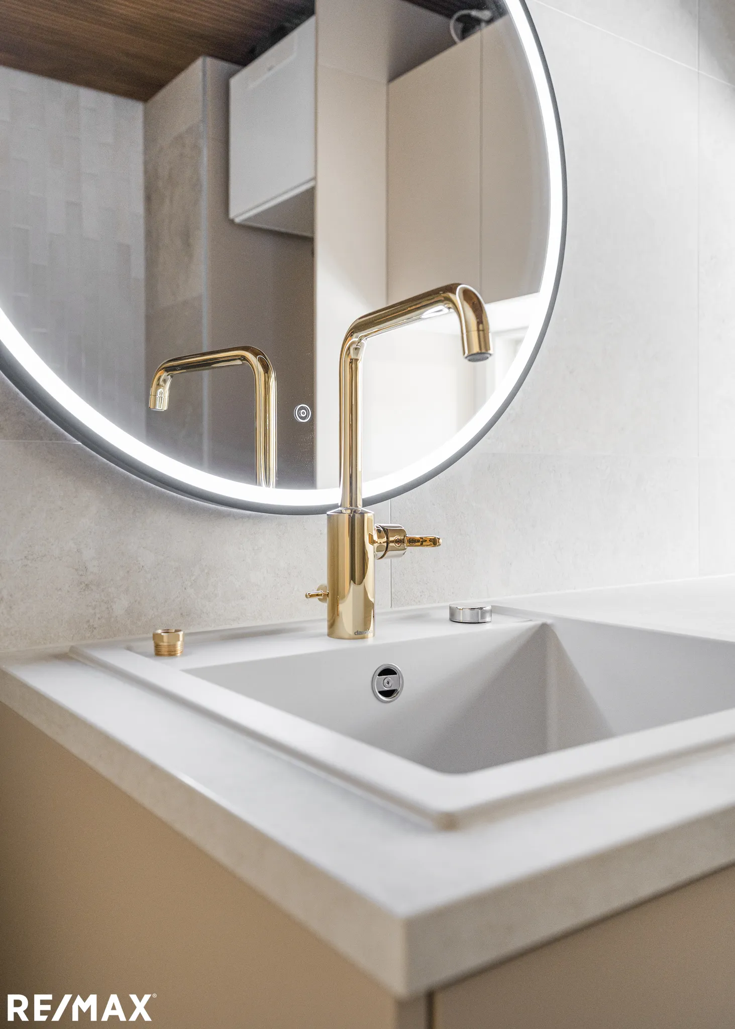 Kylpyhuoneen valkoinen pesuallas, kultainen hana ja pyöreä peili LED-valolla.