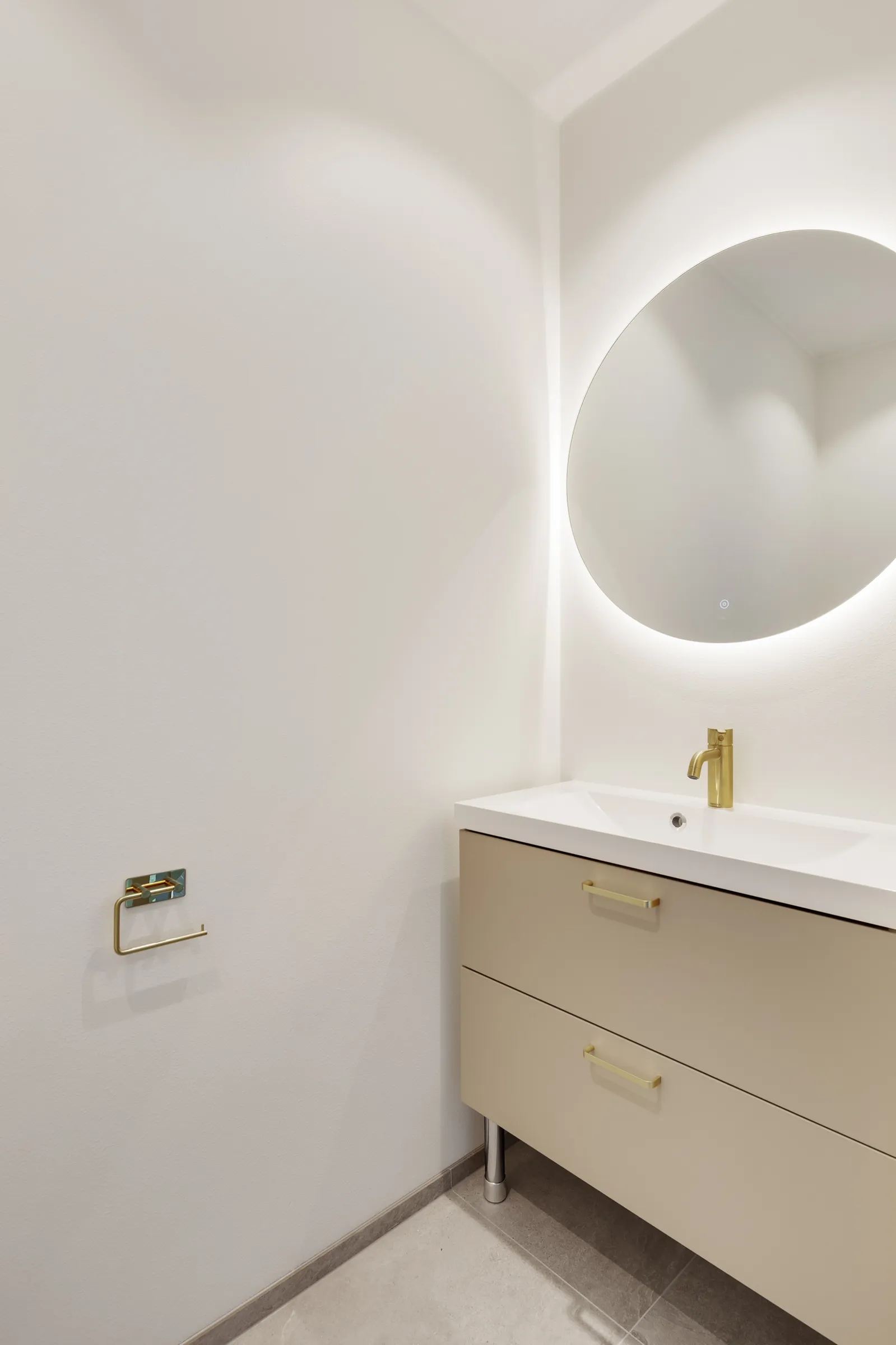 Moderni kylpyhuone pyöreällä peilillä, beigeillä laatikoilla ja kultaisella hanalla.