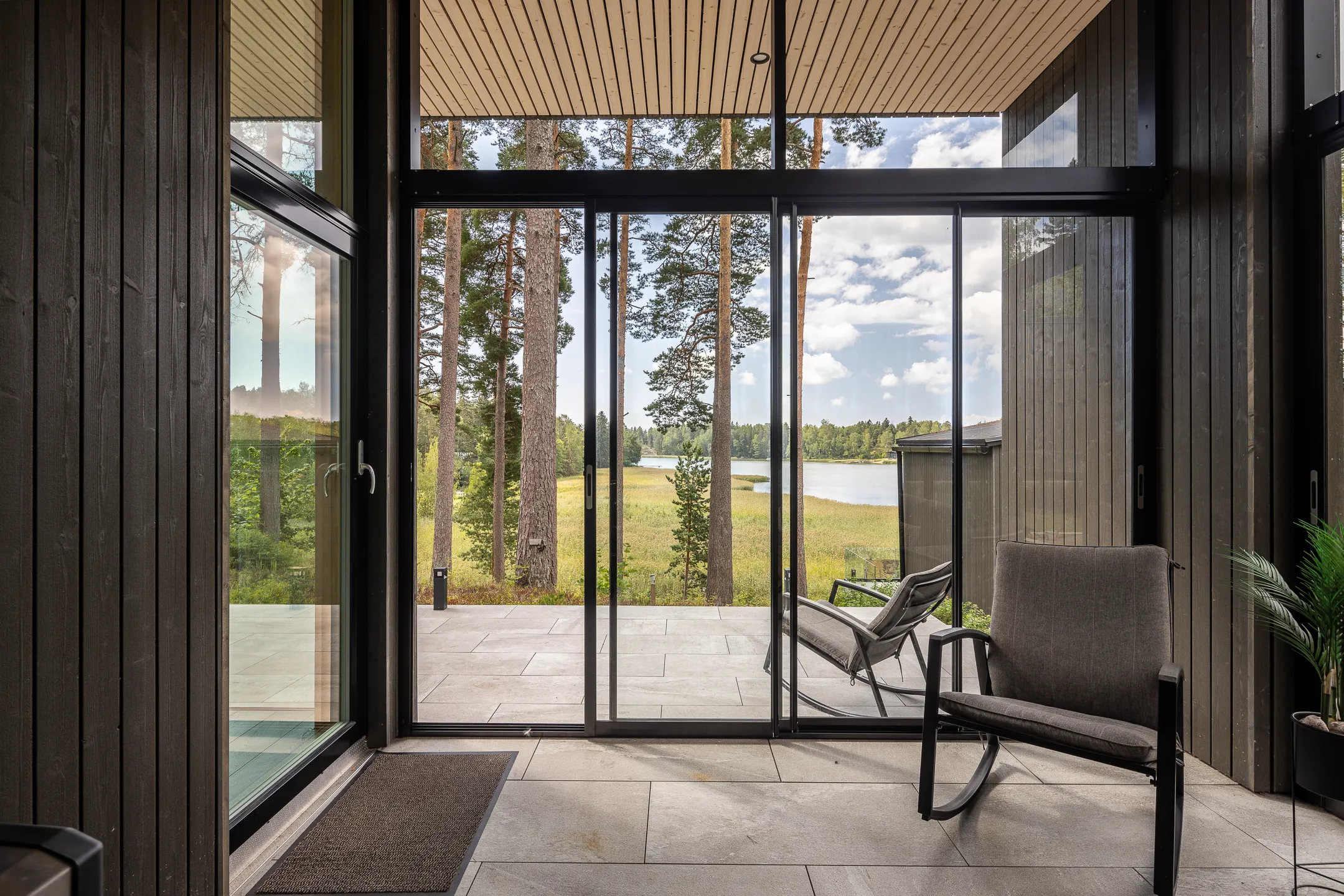 Moderni terassi, jossa on suuret lasiovet, kaksi harmaata keinutuolia ja näkymä järvelle ja metsään, valoisa ja avara tunnelma.