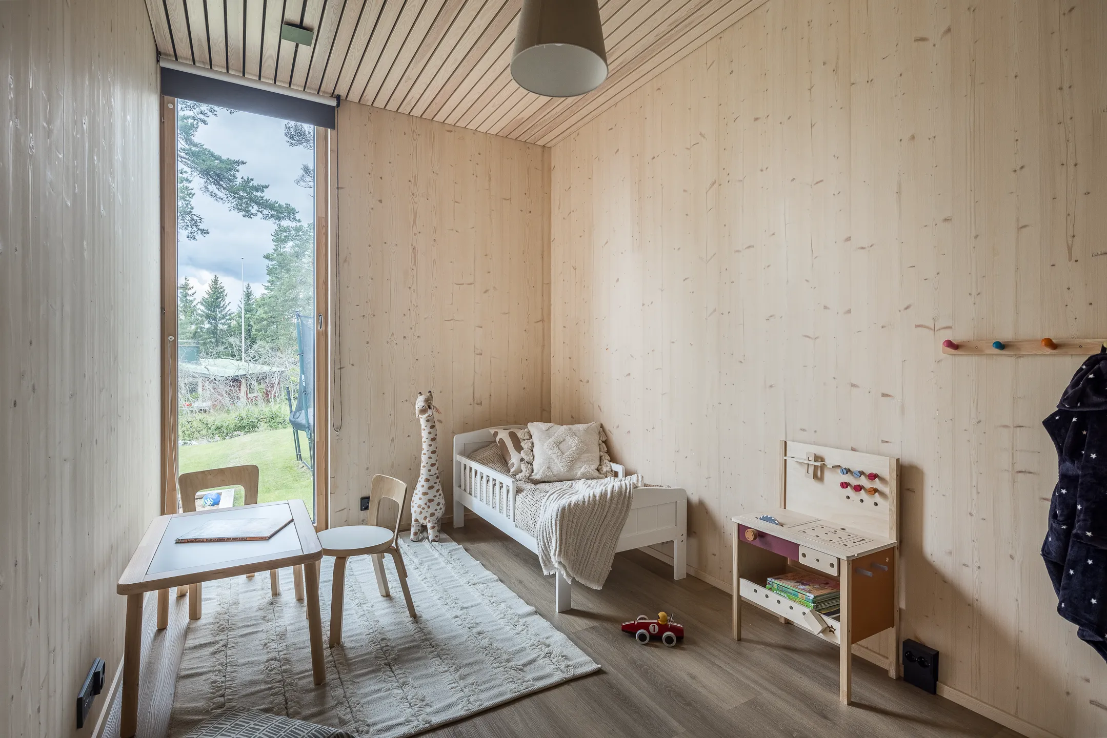 Lastenhuone, jossa on puupanelointi, sänky, pöytä, leikkityöpöytä ja ikkuna, jossa näkyy ulkona oleva maisema. Huoneessa on vaalea värimaailma ja luonnonvalo.