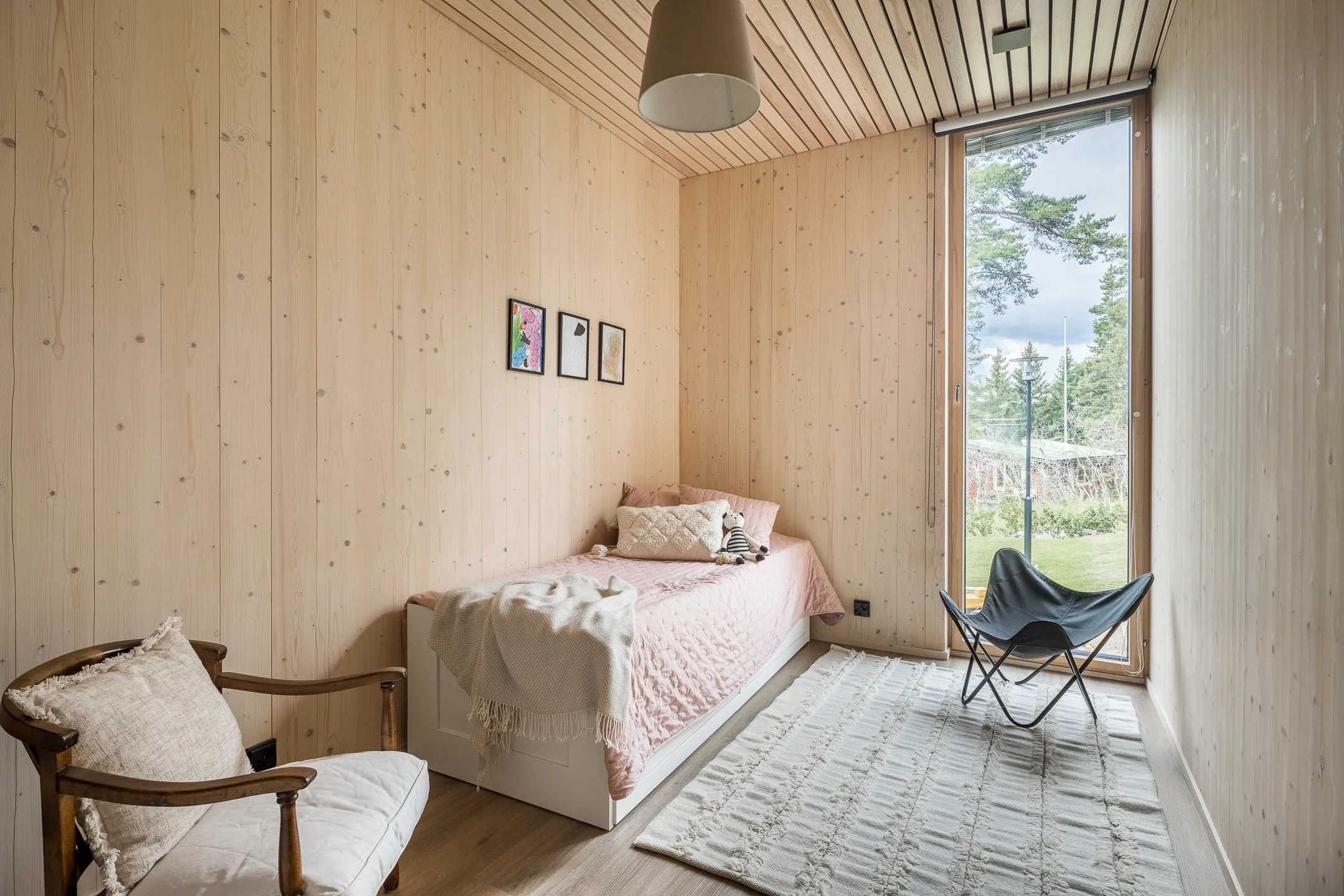 Vaaleassa makuuhuoneessa on puupaneloidut seinät, sänky vaaleanpunaisella päiväpeitteellä, nojatuoli ja ikkuna, josta näkyy ulos.