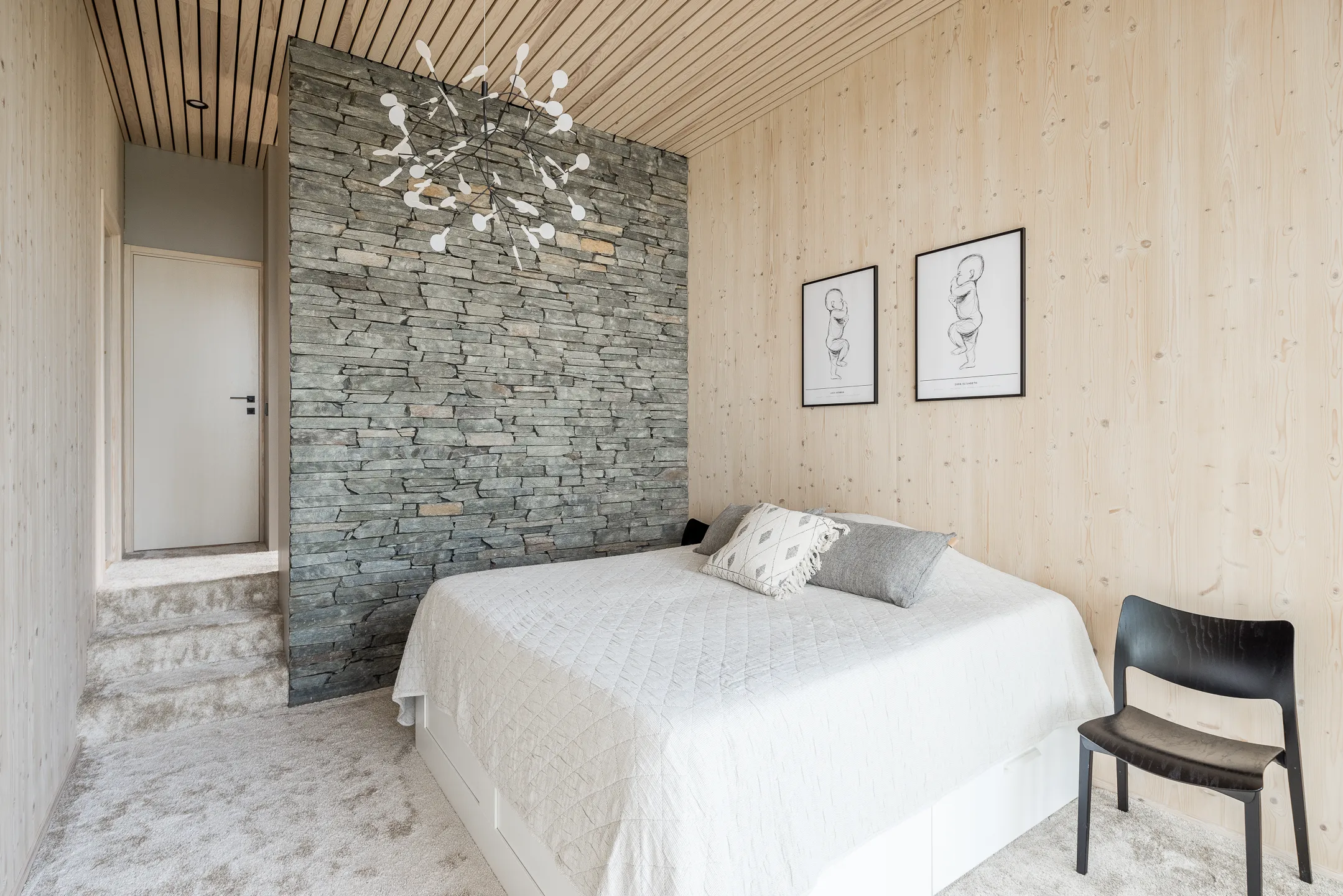Makuuhuoneessa on kiviseinä, puupanelointi, valkoinen sänky, musta tuoli ja kaksi kehystettyä taideteosta. Huoneessa on myös moderni kattokruunu ja pehmeä matto.