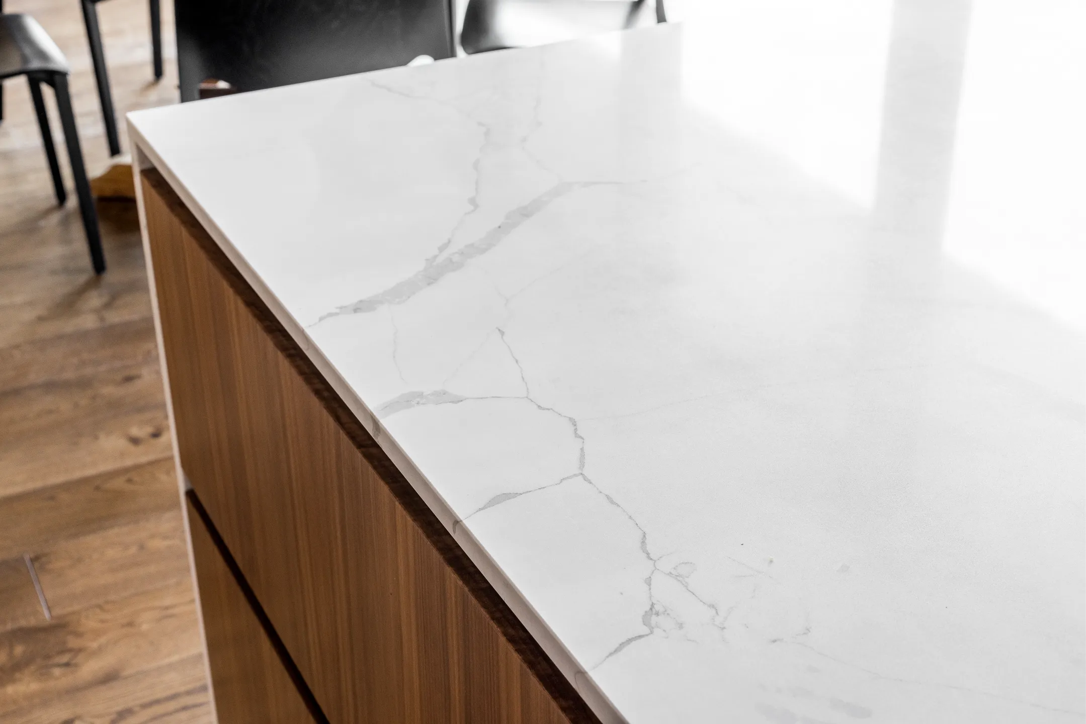 Lähikuva marmoripintaisesta keittiösaarekkeesta, jossa on puuviiluinen etuosa ja taustalla puulattia ja tuolit.