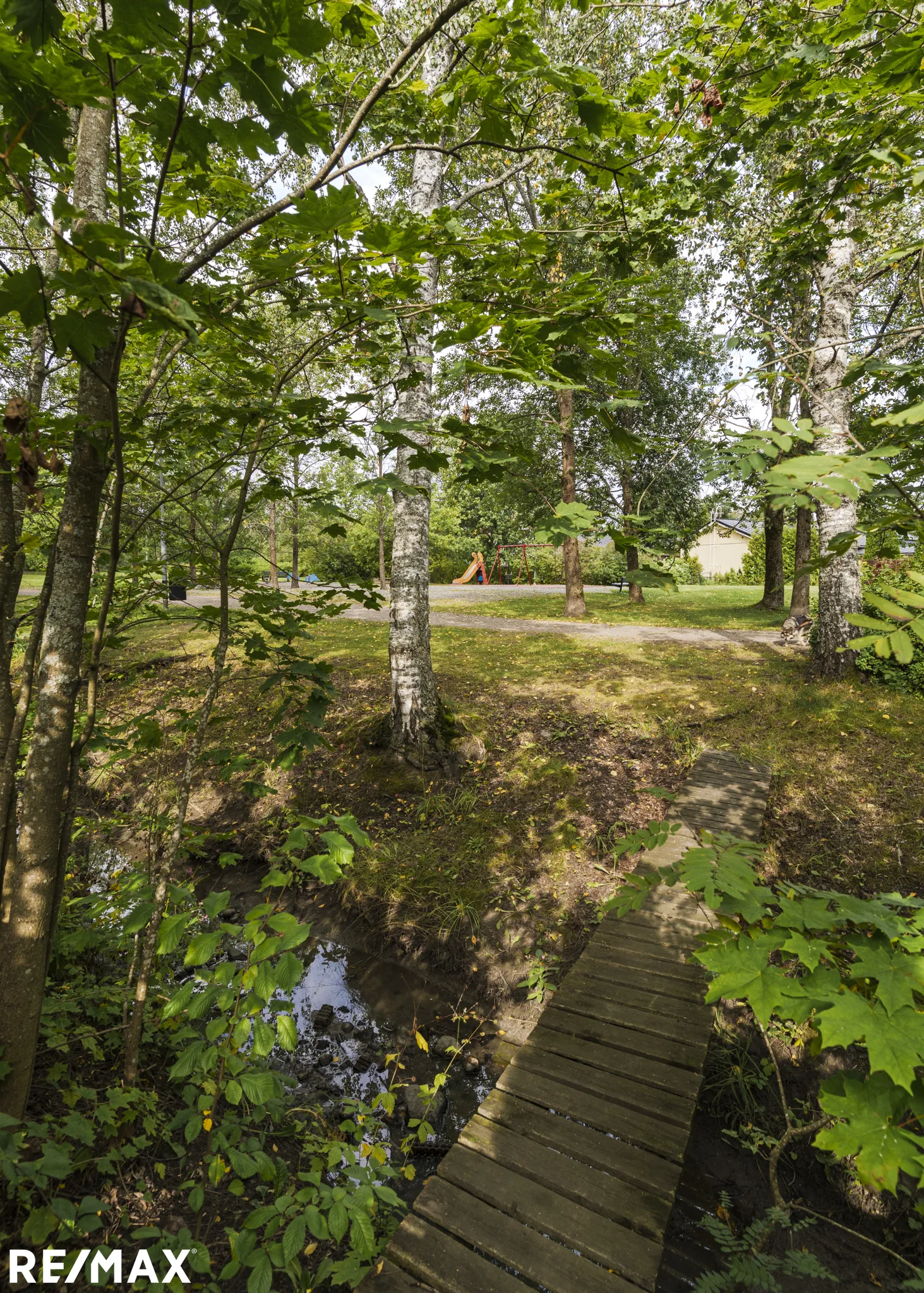 Metsäinen maisema, jossa on pieni puusilta ojan yli, vihreitä puita ja lehtiä, sekä RE/MAX-logo alareunassa.