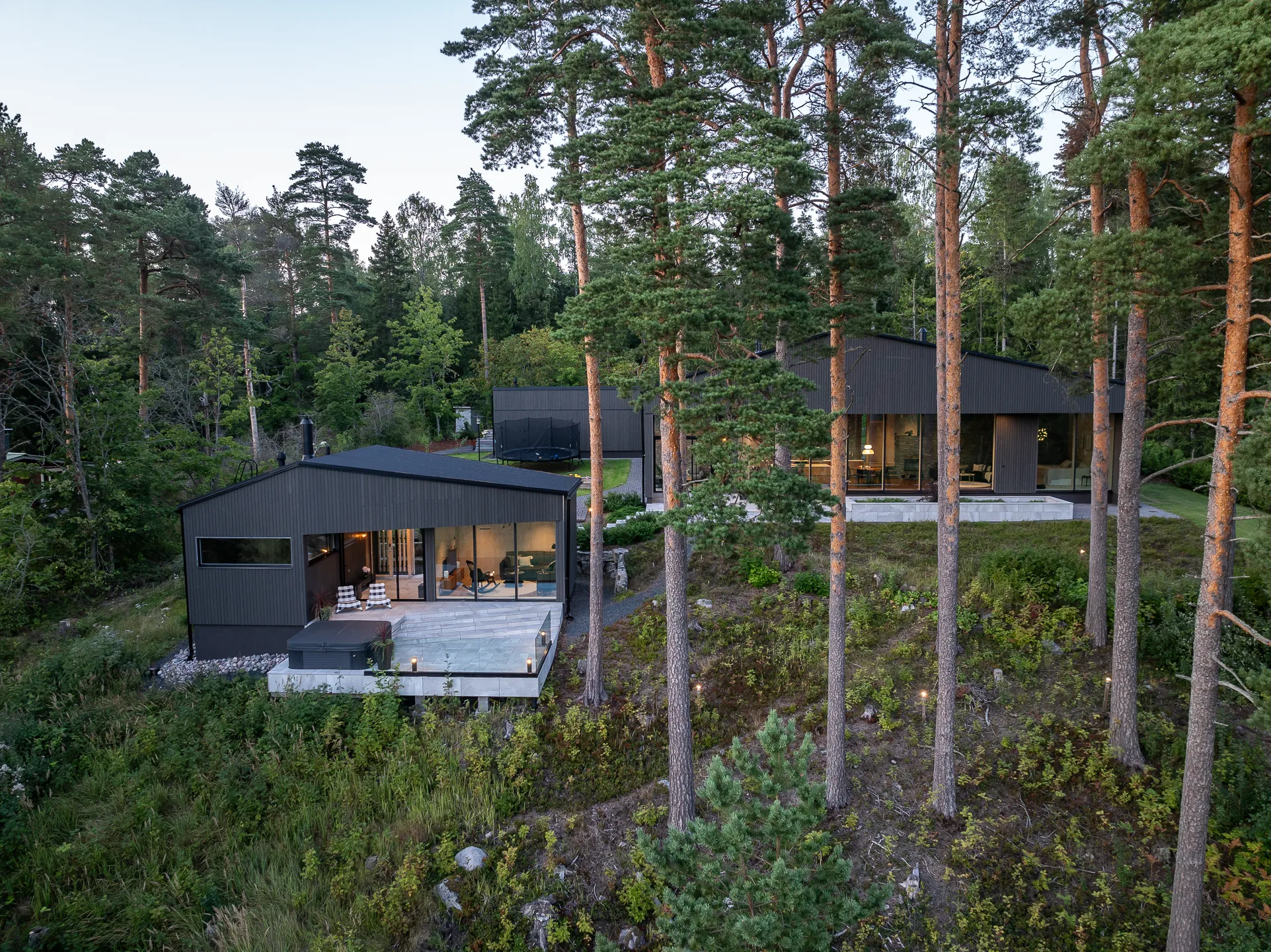 Moderni, tumma puutalo metsäisellä rinteellä, jossa on suuret ikkunat ja terassi, valaistu ulkovalaistuksella.