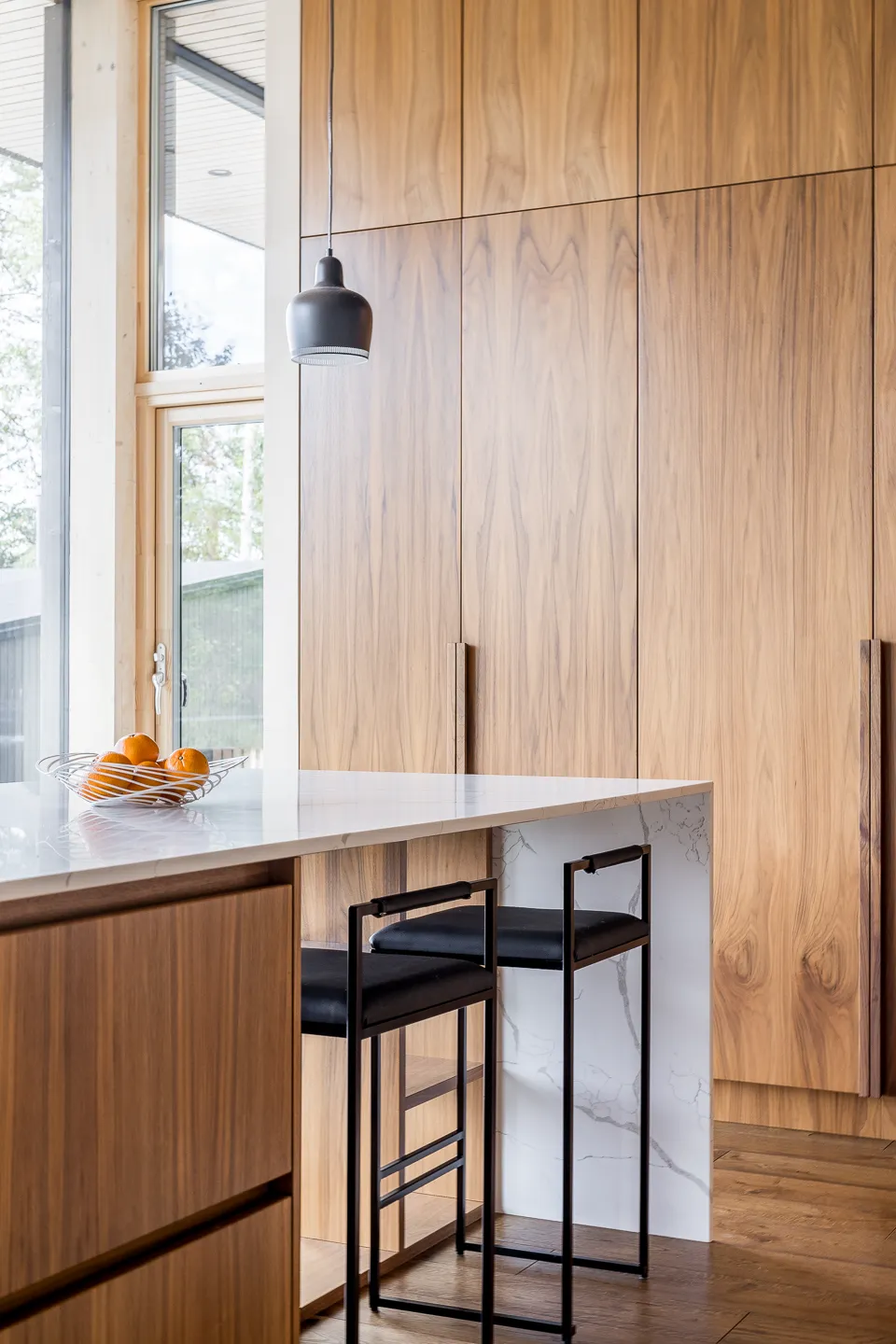 Moderni keittiö, jossa on puukaapit, marmoritaso, kaksi mustaa baarijakkaraa ja ikkunasta tuleva luonnonvalo.