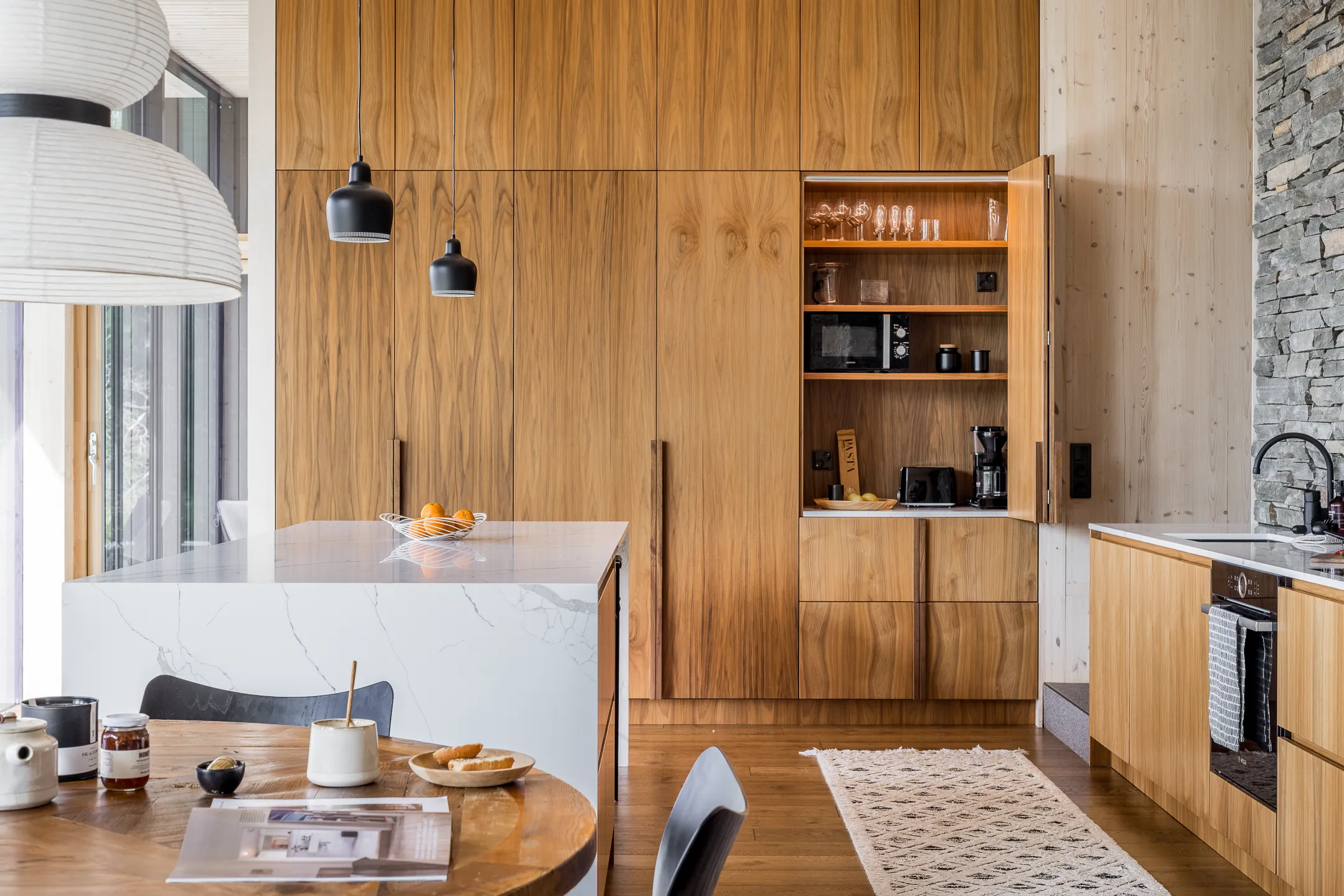 Moderni keittiö, jossa on puukaapit, marmorinen saareke, mustat riippuvalaisimet ja pyöreä puinen ruokapöytä. Avohyllyssä on lasitavaraa ja mikroaaltouuni.