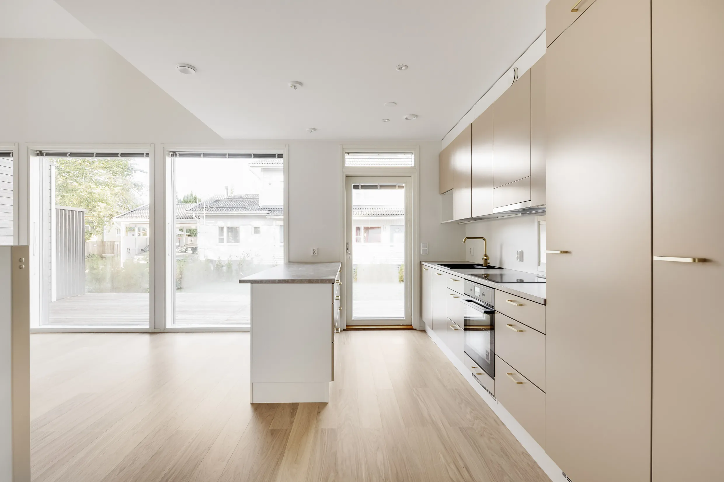 Valoisa ja moderni keittiö, jossa on vaaleat puulattiat, beigejä kaappeja kultaisilla vetimillä ja valkoinen keittiösaareke. Suuret ikkunat tuovat luonnonvaloa tilaan.