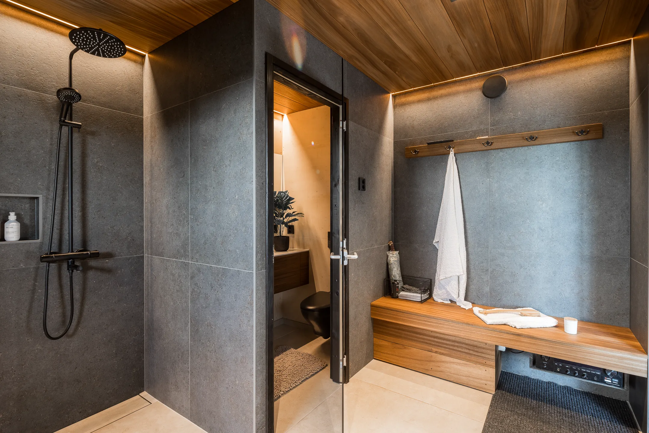 Moderni kylpyhuone, jossa on harmaat laatat, puinen katto, musta suihku, puinen penkki ja avoin ovi pienempään wc-tilaan.