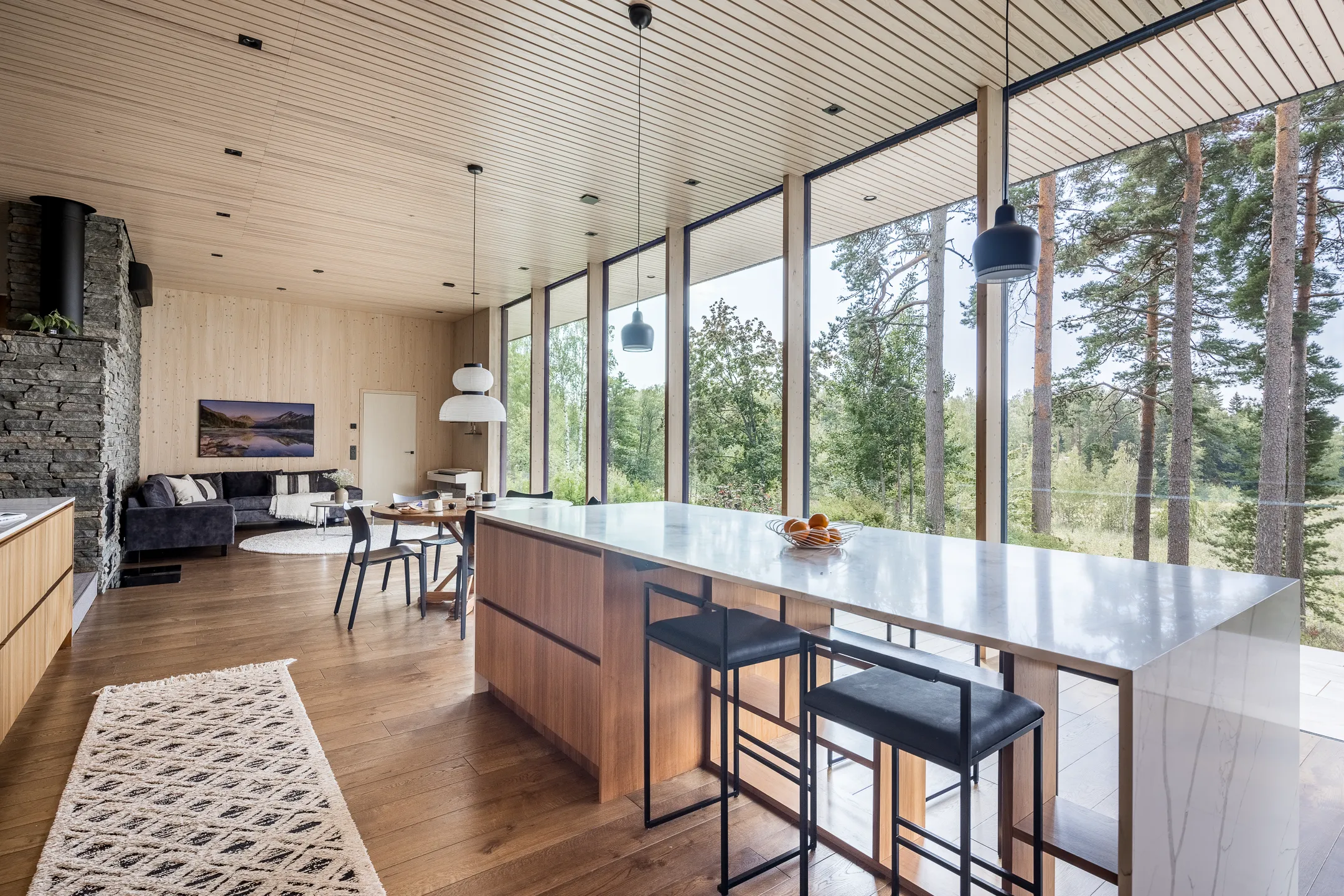 Moderni olohuone, jossa on puinen sisustus, suuret ikkunat, keittiösaareke, kivitakka, harmaa sohva ja geometrinen matto. Valoisa ja avara tila.