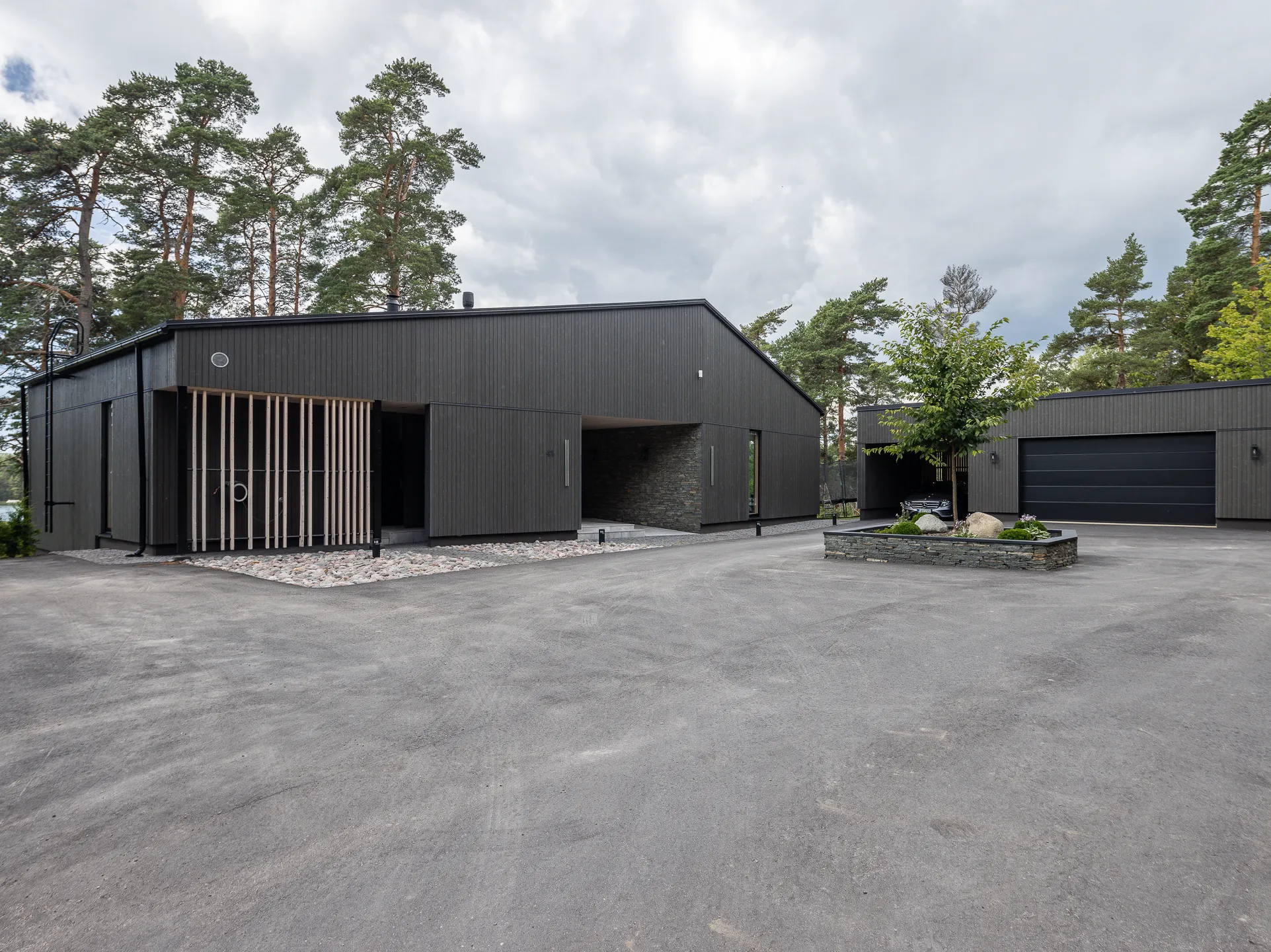 Moderni, tumma puutalo, jossa on harmaa ajotie, puita ja pilvinen taivas.