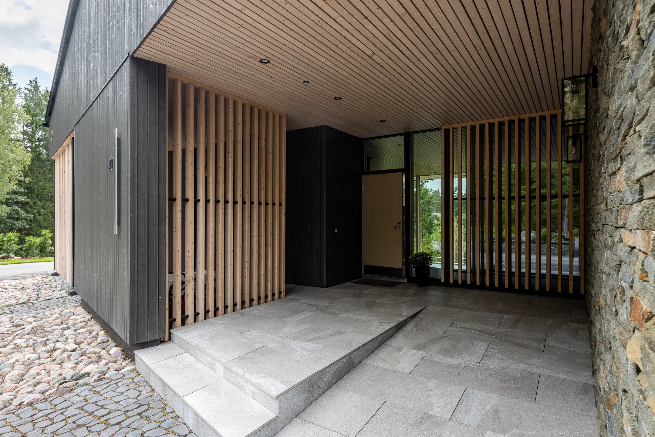 Modernin talon sisäänkäynti kiviseinällä, puurimoituksella ja harmaalla puuverhoilulla, jossa on laatoitettu lattia ja puinen kattopaneeli.