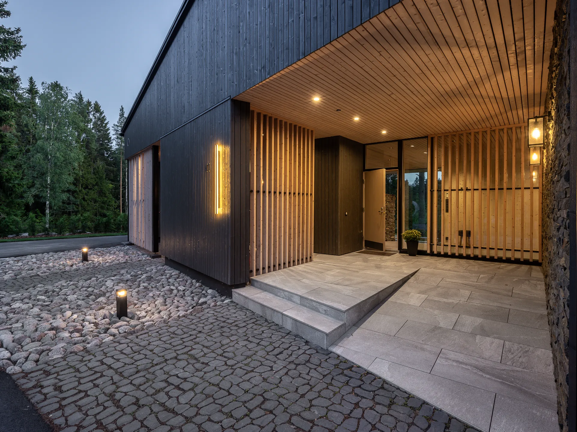 Modernin talon sisäänkäynti hämärässä, jossa on puuverhoilu, kivipolku ja ulkovalaisimet. Tumma puuverhous ja vaaleat puurimat luovat kontrastin.