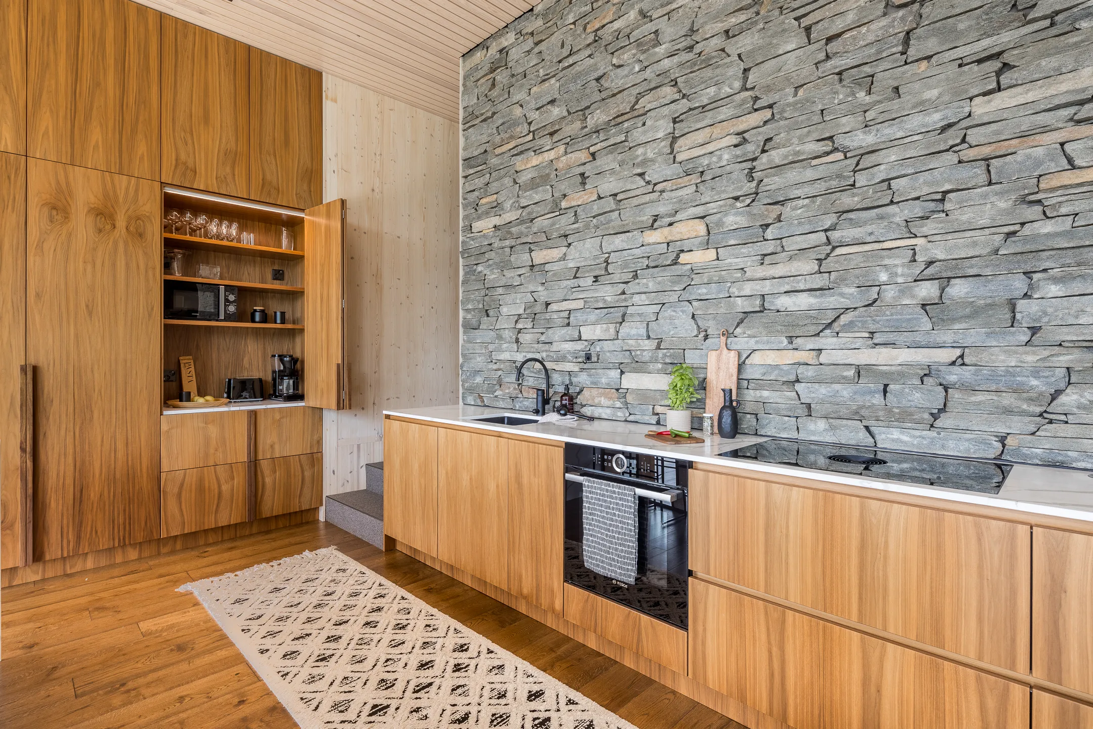 Moderni keittiö, jossa on puukaapit, kiviseinä ja valkoinen työtaso. Lämmin valaistus korostaa puun ja kiven luonnollisia sävyjä.