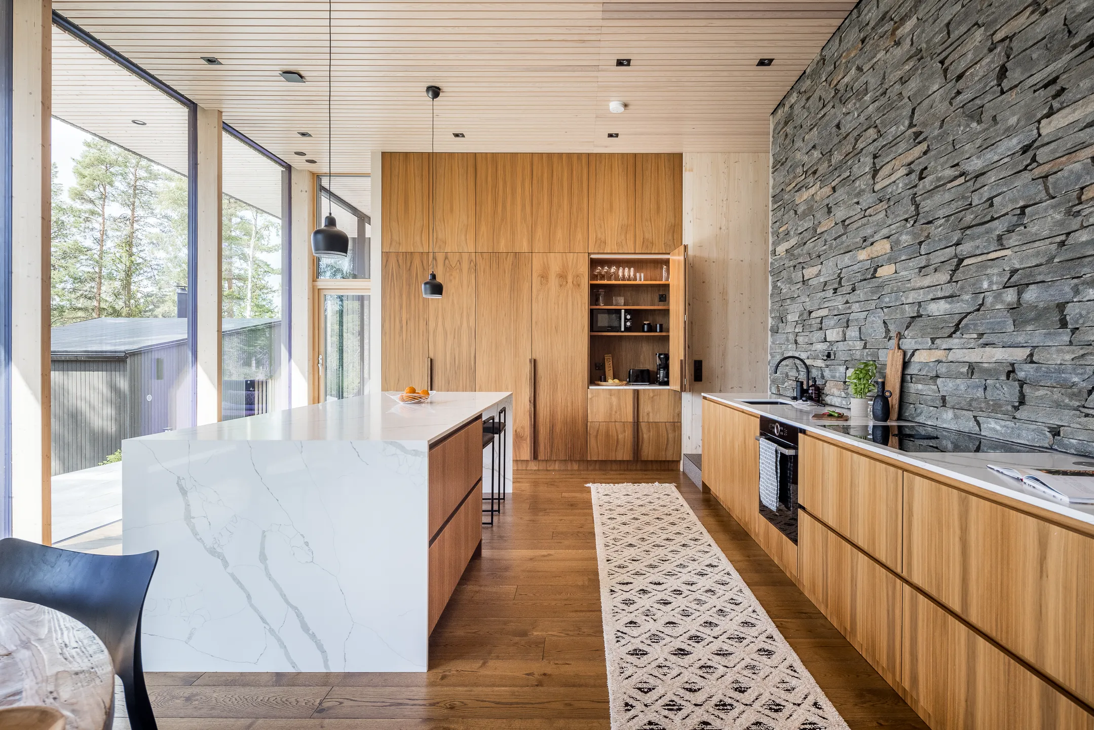 Moderni keittiö, jossa on puuta, kiveä, valkoinen saareke ja pitkä matto puulattialla. Suuret ikkunat tuovat luonnonvaloa.